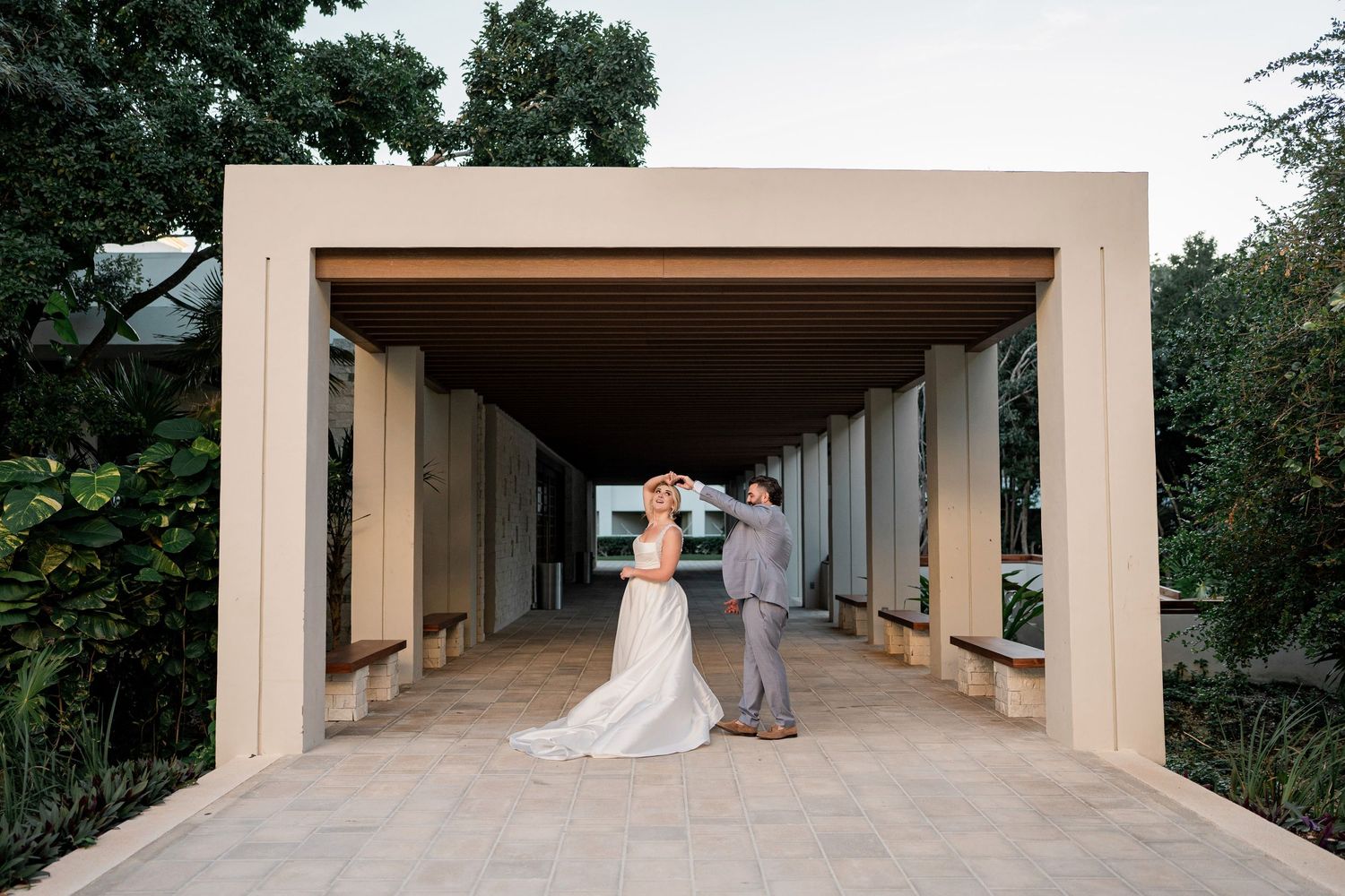 Bride and groom share a romantic moment under a modern pavilion surrounded by lush greenery at an outdoor wedding venue.