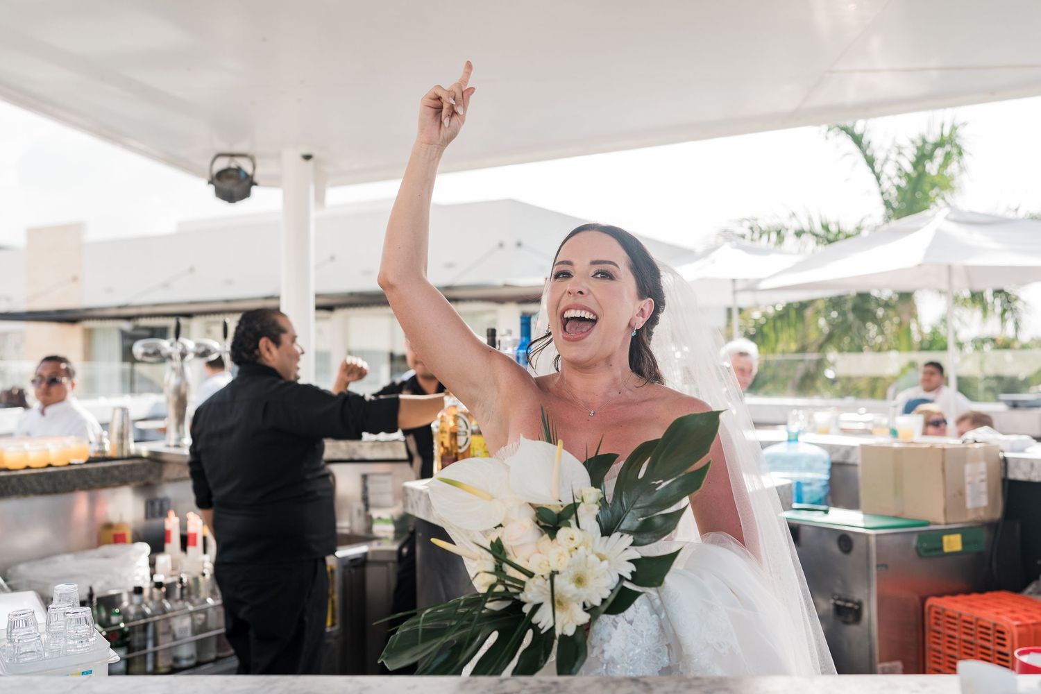 Joyful bride in white gown raises hand holding bouquet of white flowers at outdoor wedding reception bar.