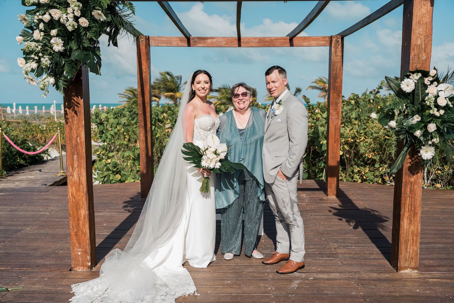 Bride and groom pose with officiant under wooden floral arch at outdoor beach wedding ceremony.