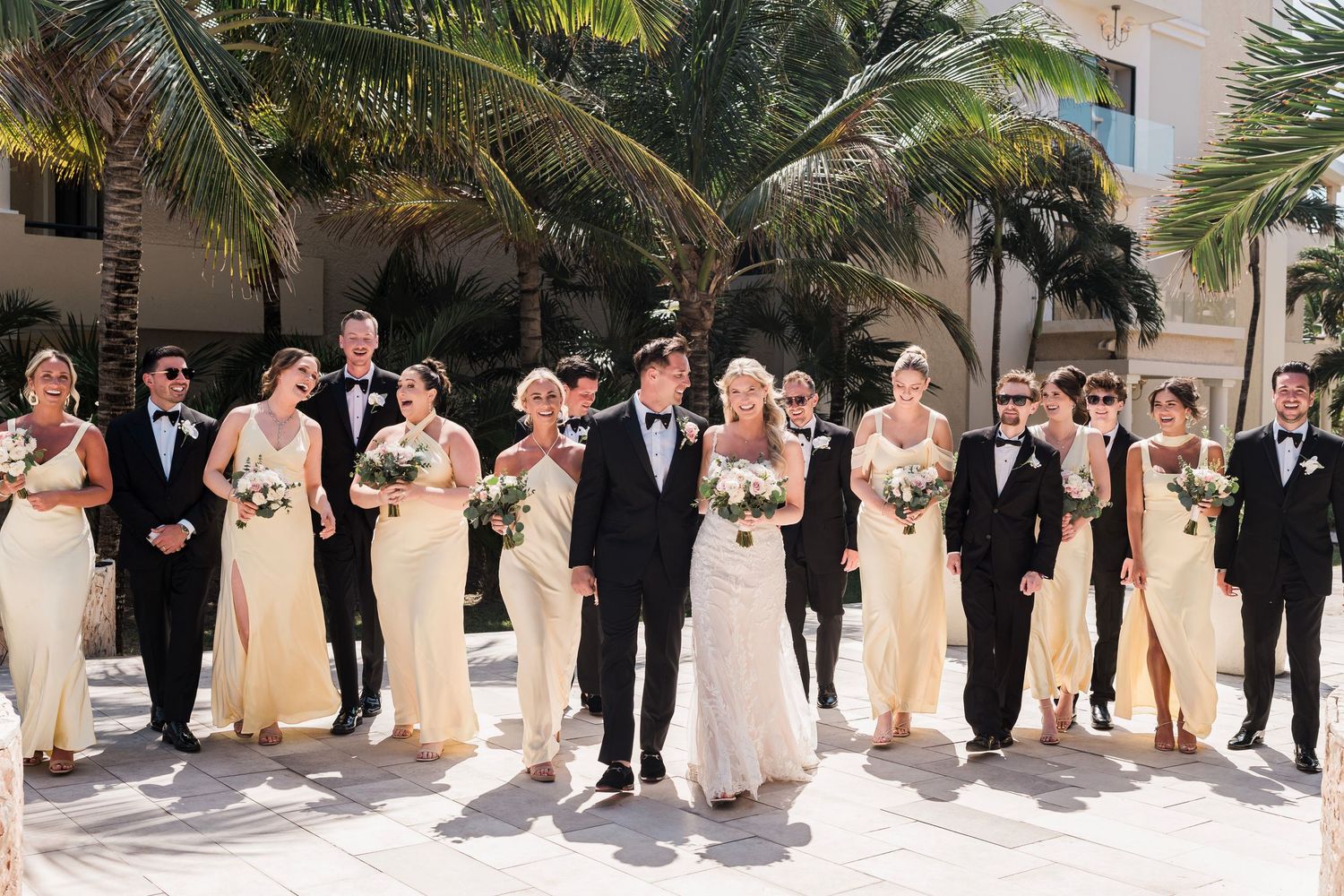 Wedding party walks outdoors near palm trees, bridesmaids in champagne gowns and groomsmen in black tuxedos flanking the bride and groom.