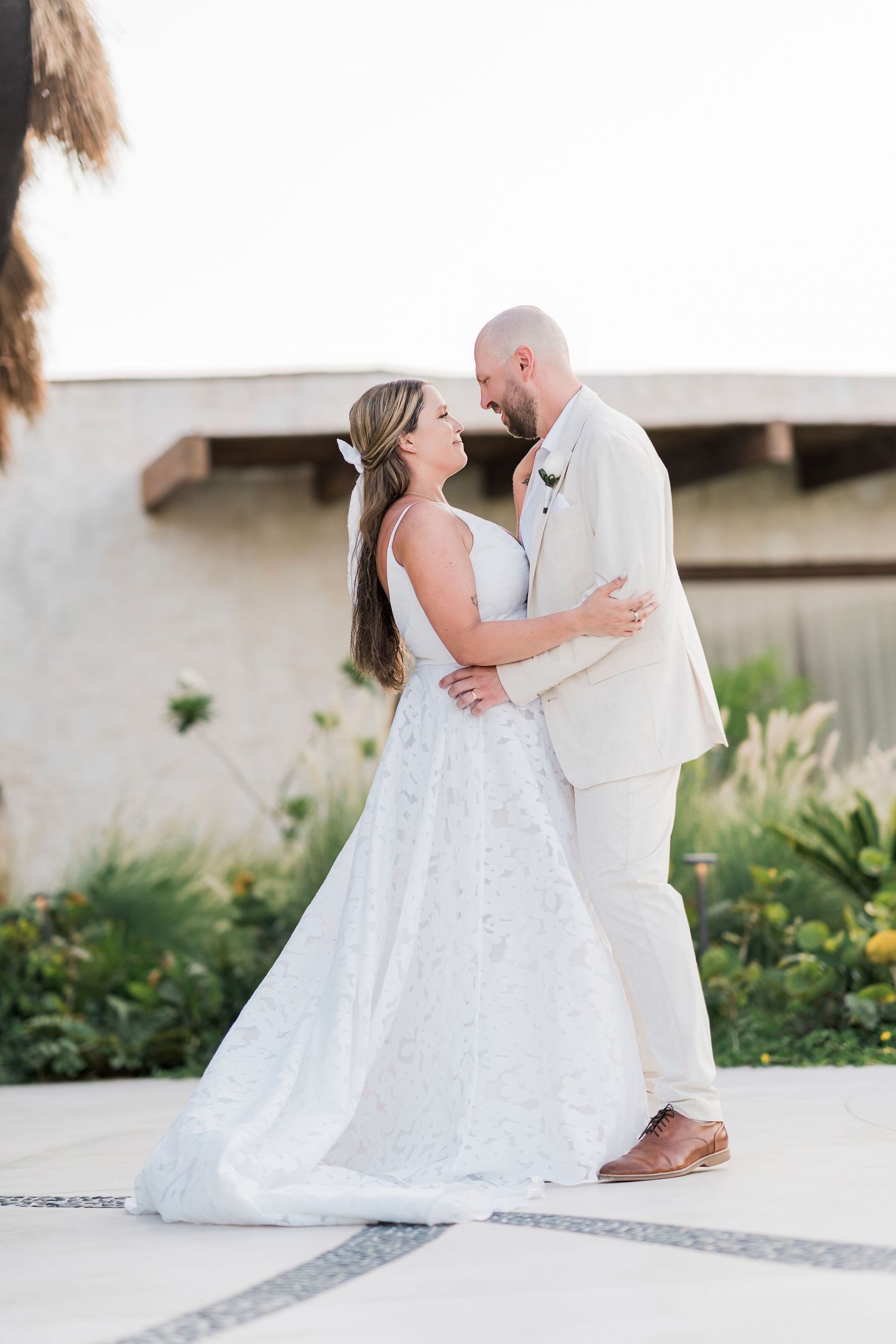 A bride in a white lace gown and groom in a beige suit share a romantic embrace outdoors on their wedding day.
