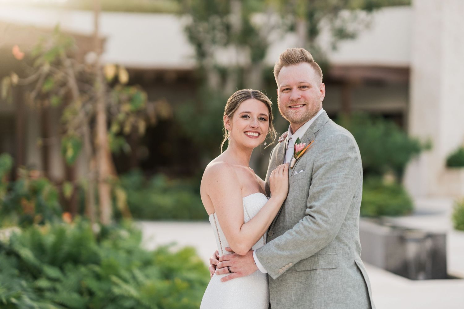 A smiling bride in a white strapless gown and groom in a gray suit pose together outdoors at their wedding.