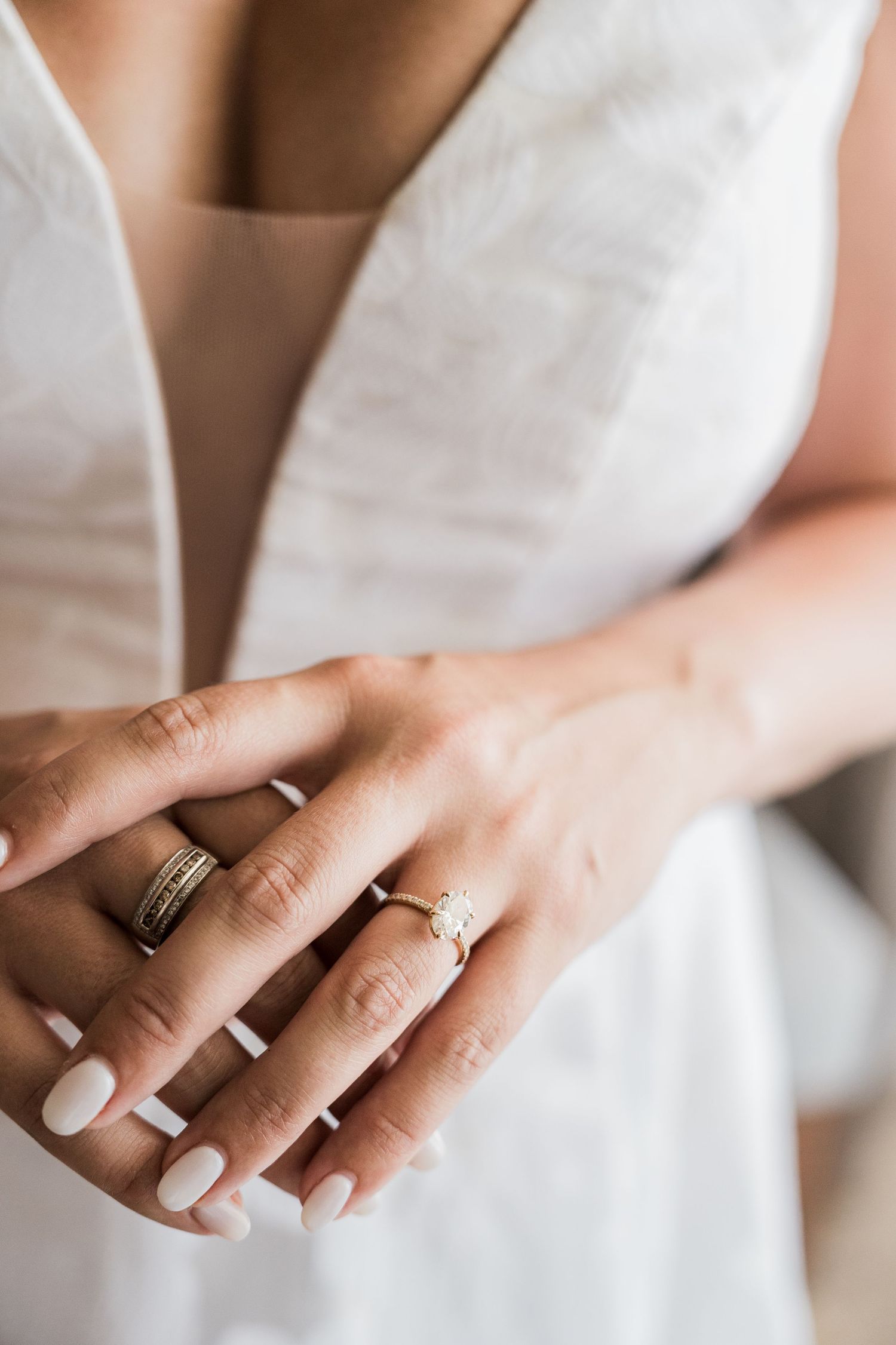 Bride wearing diamond engagement ring and gold band with white manicure, hands clasped over white wedding dress.