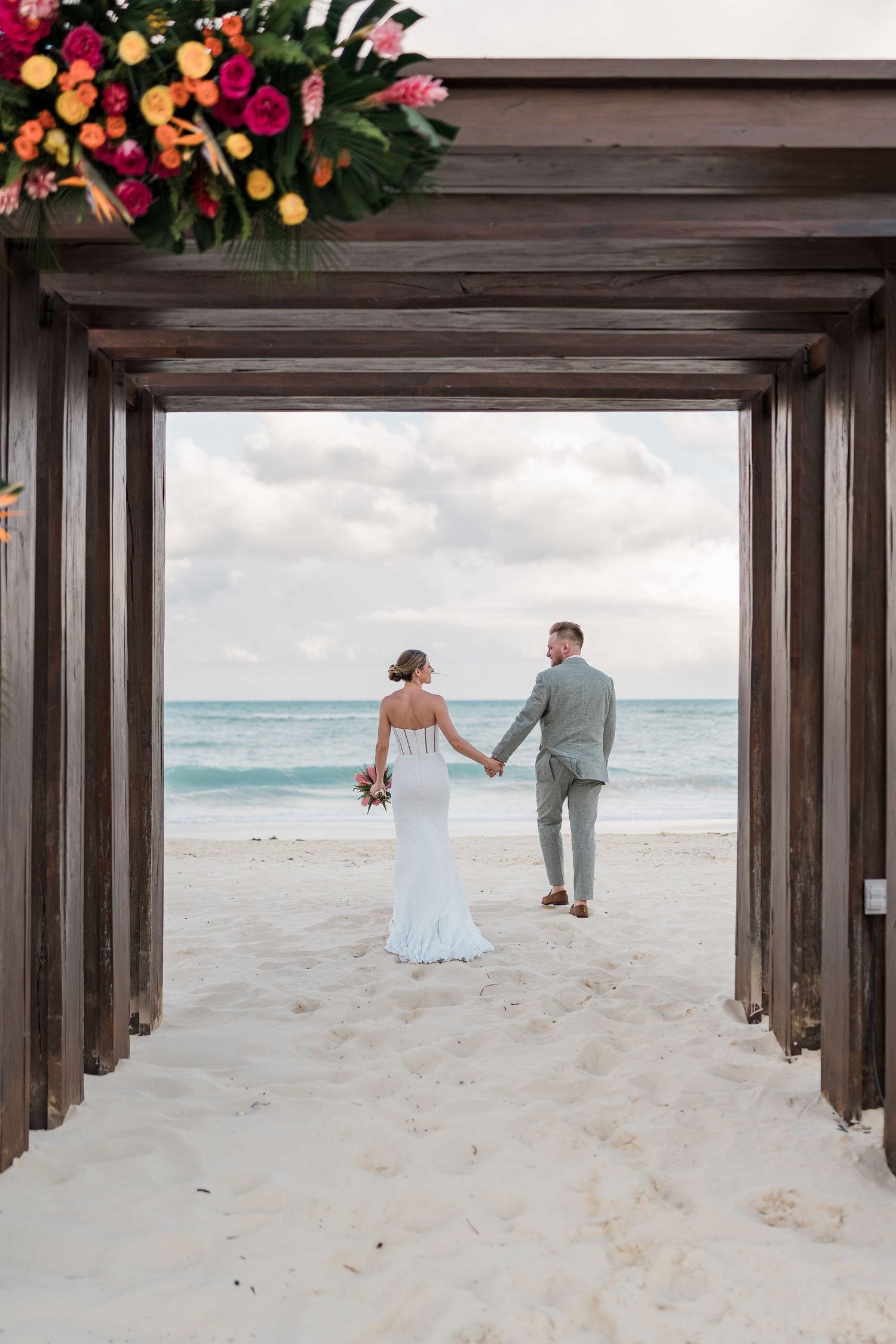 Bride and groom holding hands on a sandy beach framed by a wooden arch adorned with tropical flowers.