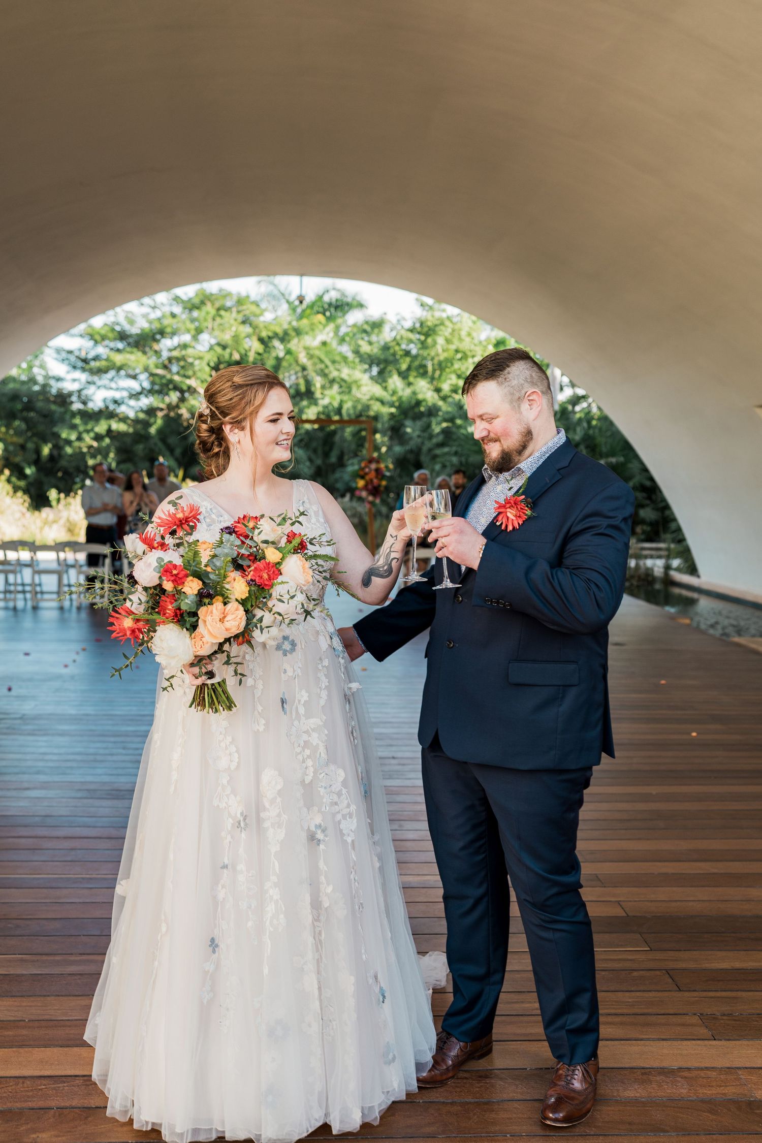Bride and groom toasting with champagne glasses, she holds a colorful bouquet, standing under an arch at their wedding venue.