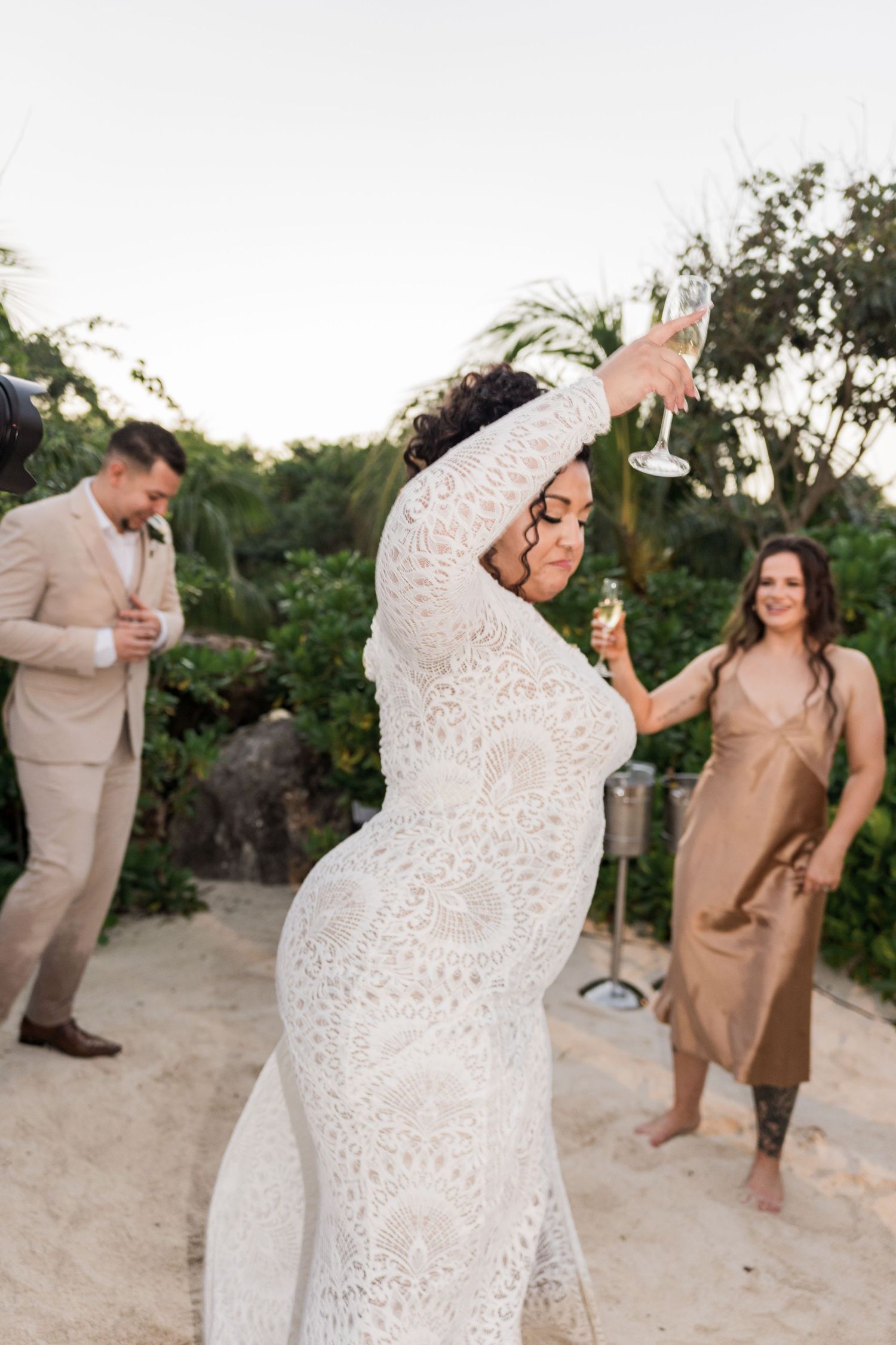 Xcaret Arte Wedding - Bride in white lace gown dancing on beach with champagne glass at outdoor wedding celebration.