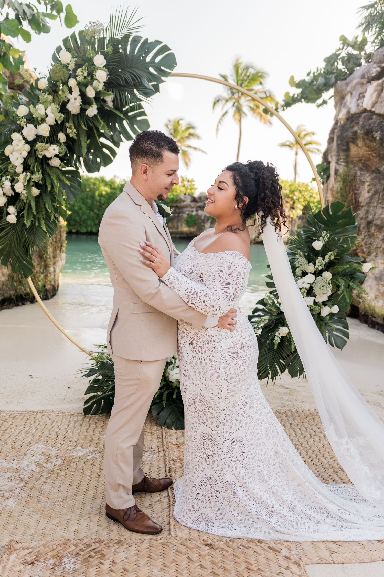 Xcaret Arte Wedding - Couple embracing at tropical beach wedding ceremony with white floral arch, palm trees, and turquoise water backdrop.