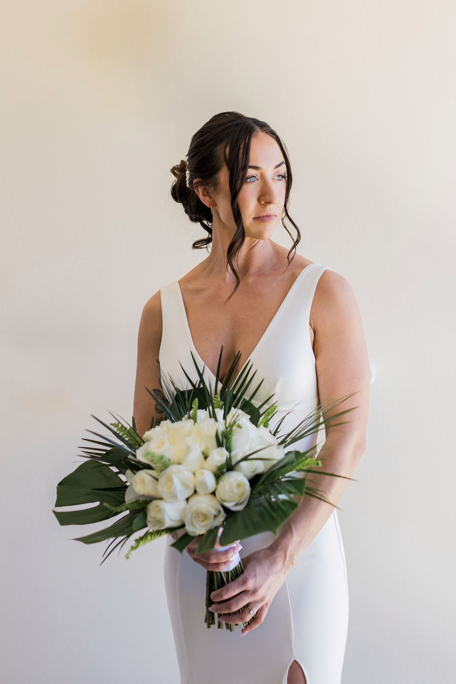 Elegant bride in white sleeveless gown holding tropical bouquet with white roses and monstera leaves against neutral background.