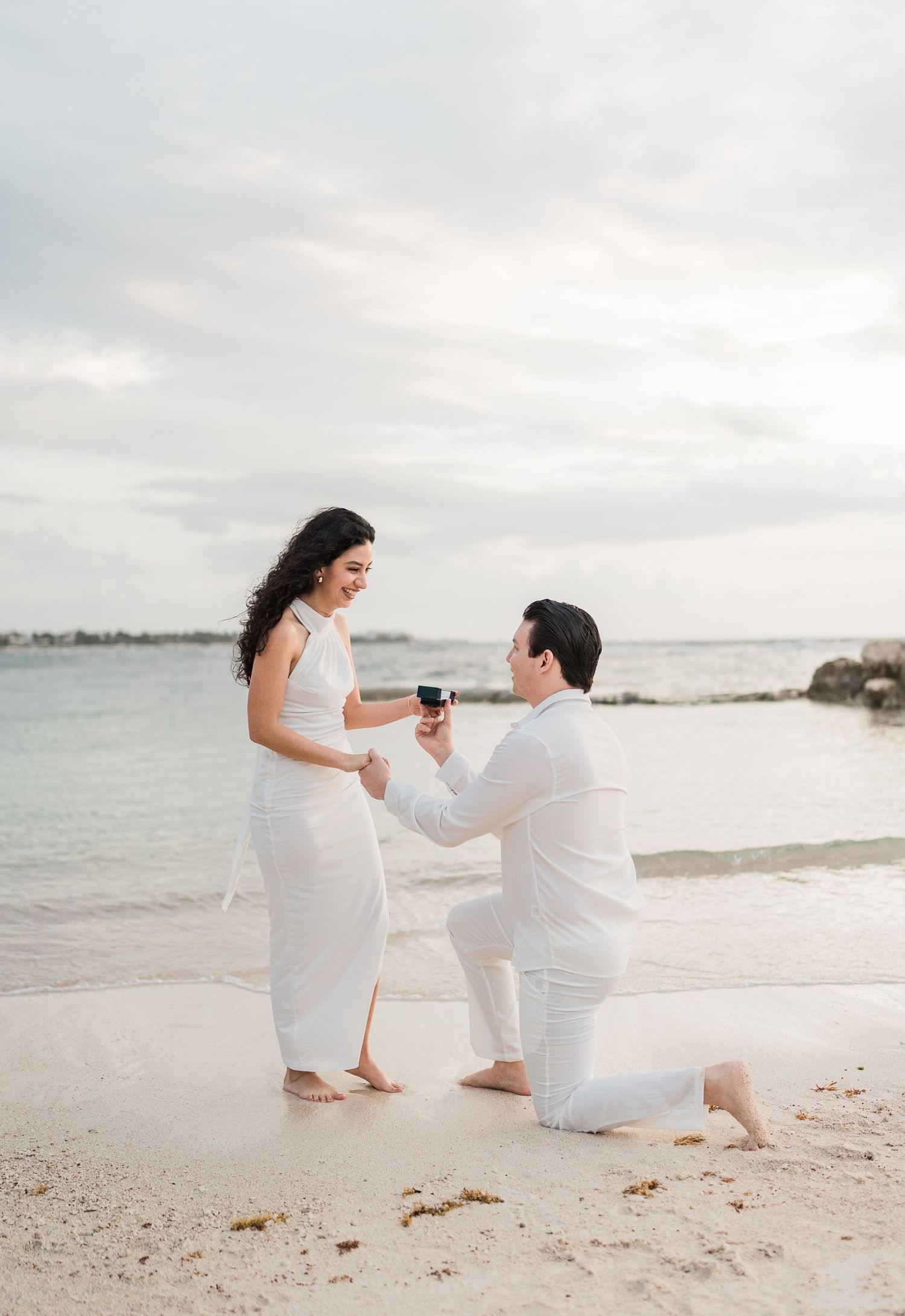 Man proposes to woman on a sandy beach, both dressed in white, with ocean waves in the background.