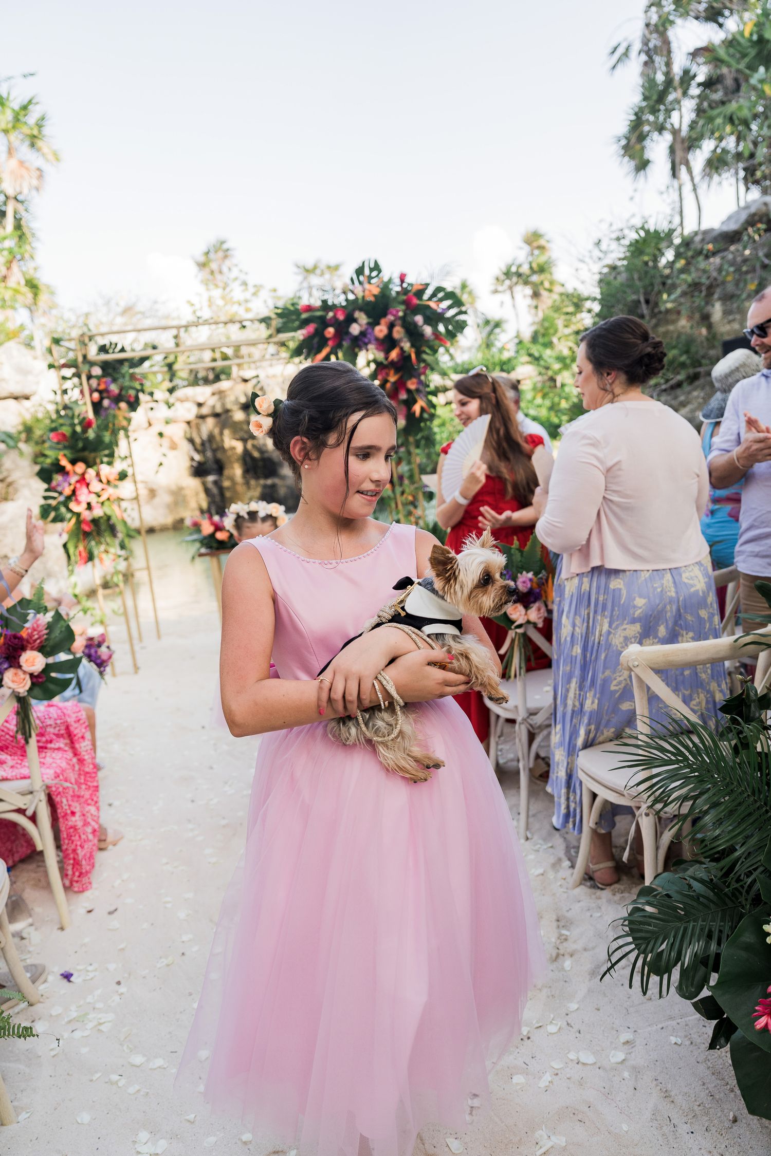 Girl in pink tulle dress holds a small Yorkshire Terrier in formal attire at an outdoor tropical wedding ceremony.