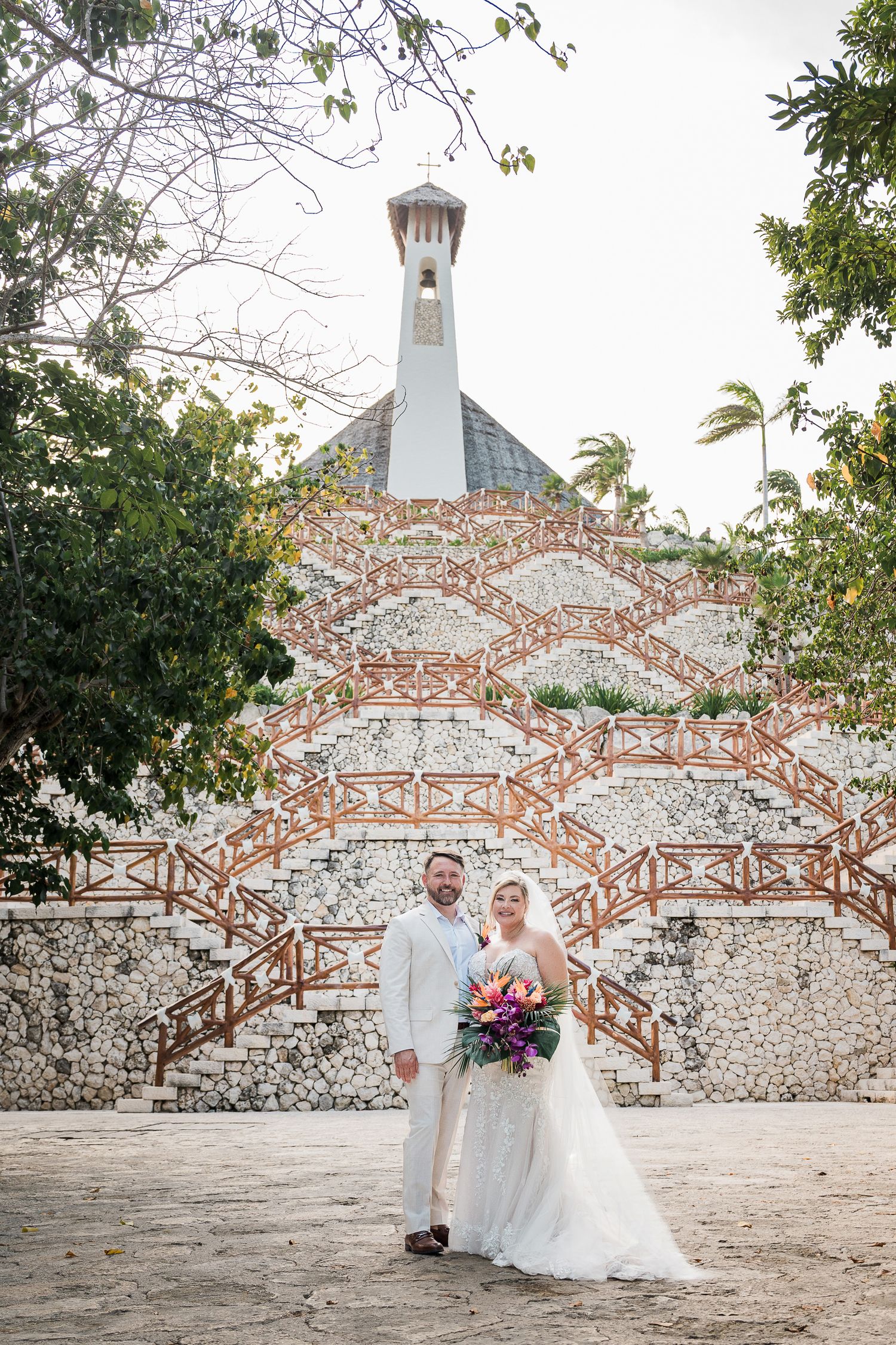 Bride and groom pose before a decorative stone chapel with brick staircases, bride holding a vibrant tropical bouquet.