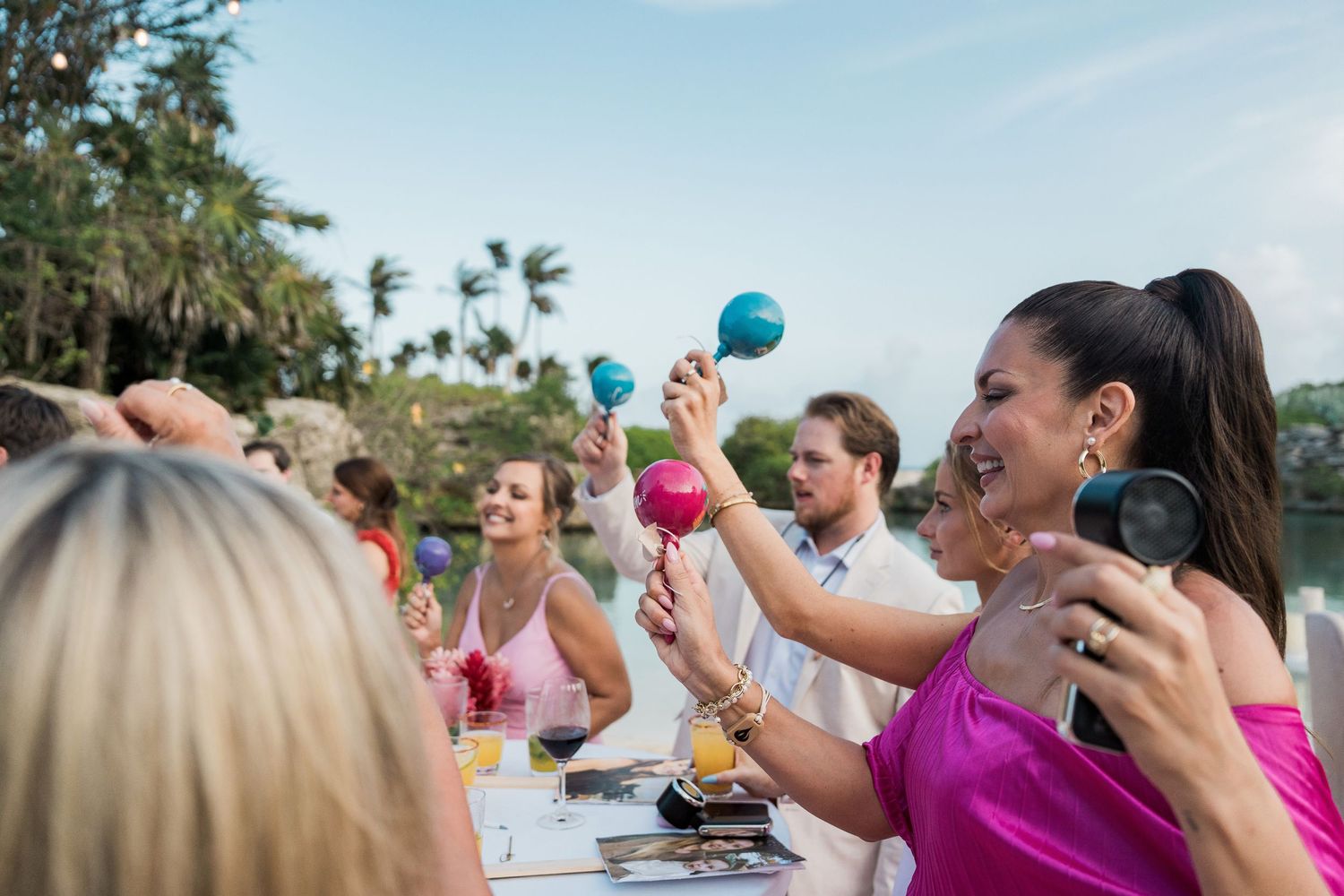 Guests at an outdoor tropical party raise colorful maracas and glasses in celebration at a festive dining table.