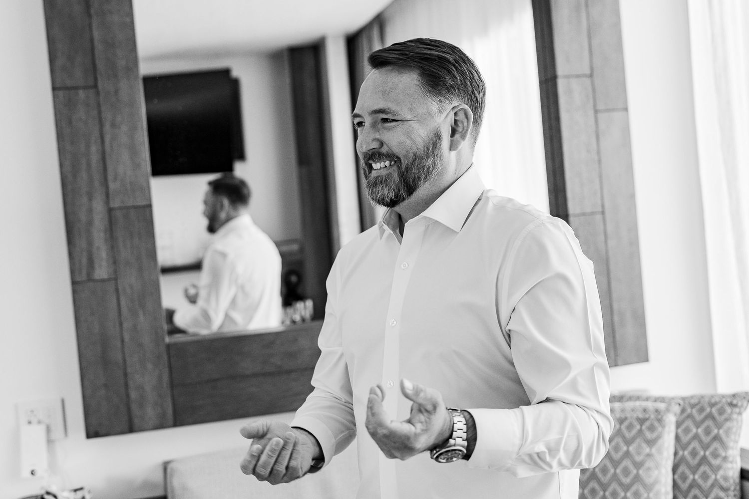 Black and white photo of a smiling bearded man in a white dress shirt, with his reflection visible in a mirror behind him.