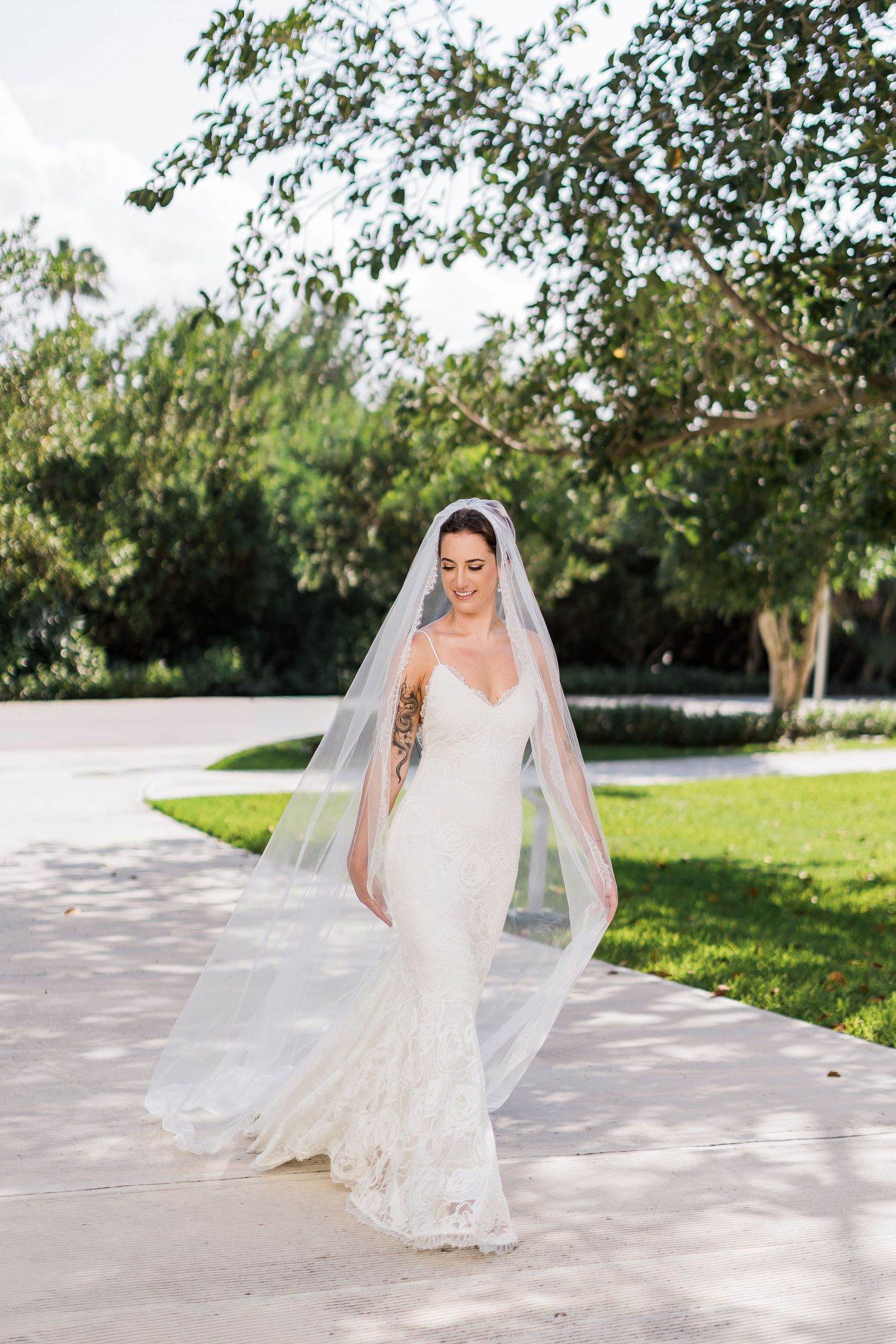 Bride in lace mermaid gown and flowing veil walks outdoors on sunny day surrounded by lush green trees.