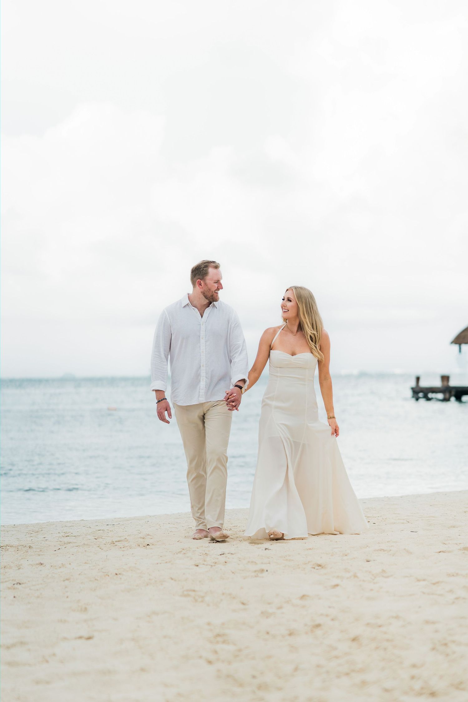 Cancun Engagement Photoshoot - A couple in white outfits holds hands while walking on a sandy beach