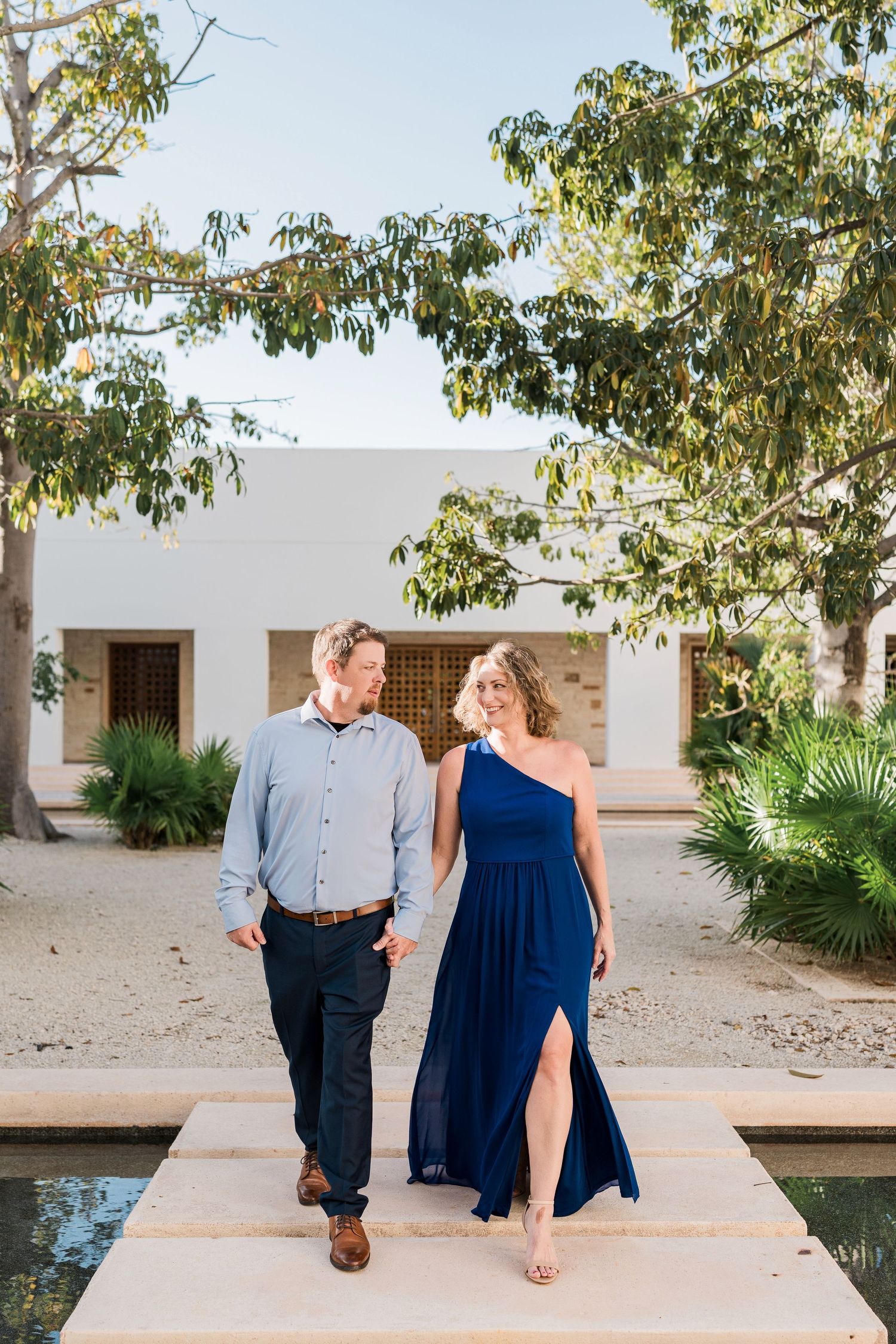 Secrets Akumal Wedding - Couple in blue dress and casual shirt walk hand-in-hand along a sunny resort pathway.