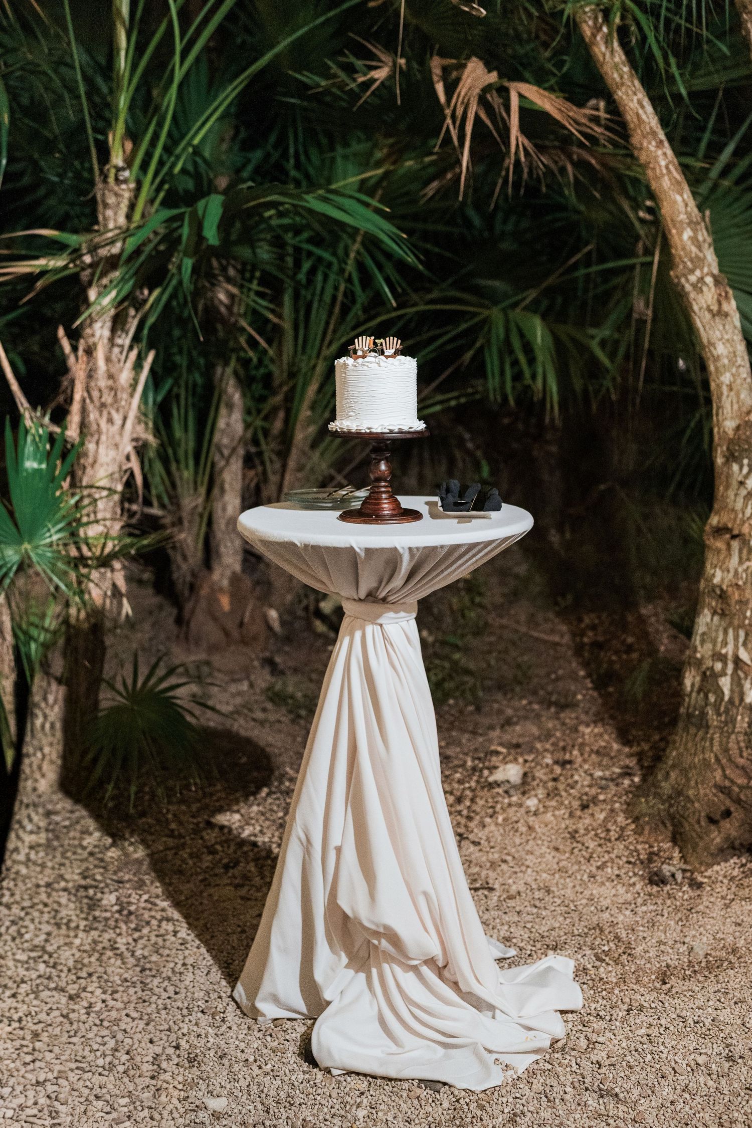 Secrets Akumal Wedding - Elegant white wedding cake on a draped cocktail table surrounded by lush tropical palms at an outdoor wedding.