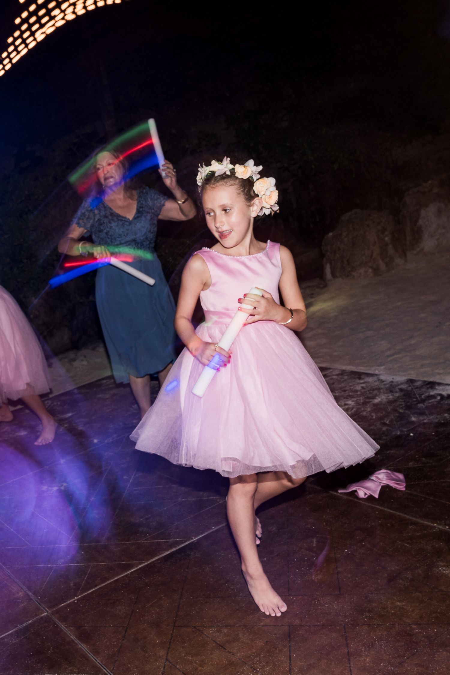 A young girl in a pink tulle dress and floral crown dances barefoot at a wedding reception with glowing light sticks.