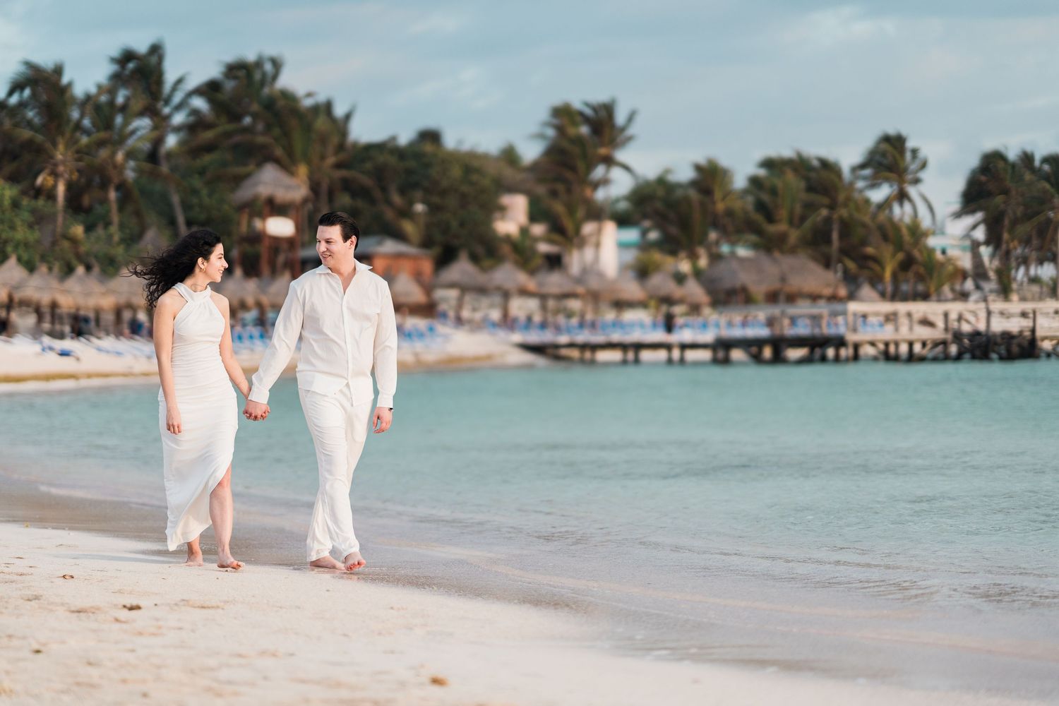 Couple in white outfits holding hands walking on a tropical beach with palm trees and resort pier in background.