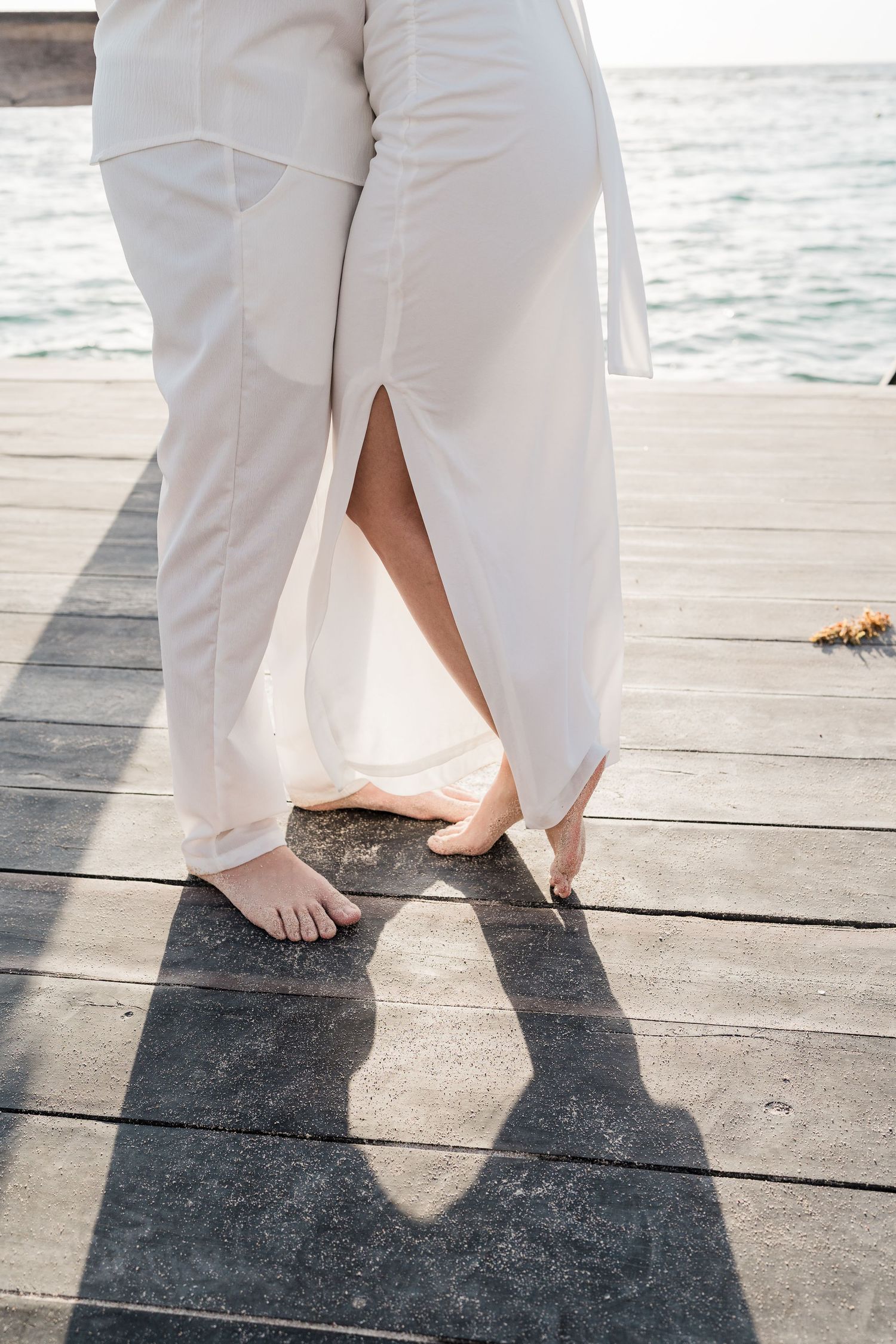 Barefoot couple in white attire standing on a wooden pier by the ocean, casting a heart-shaped shadow.