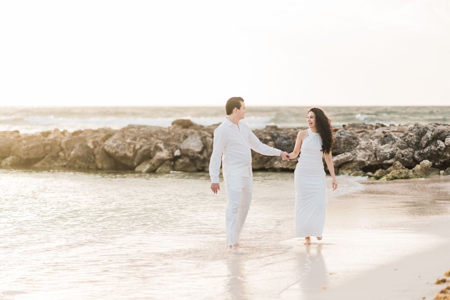 Couple in white outfits holding hands and walking on a sandy beach with rocky shoreline at sunset.