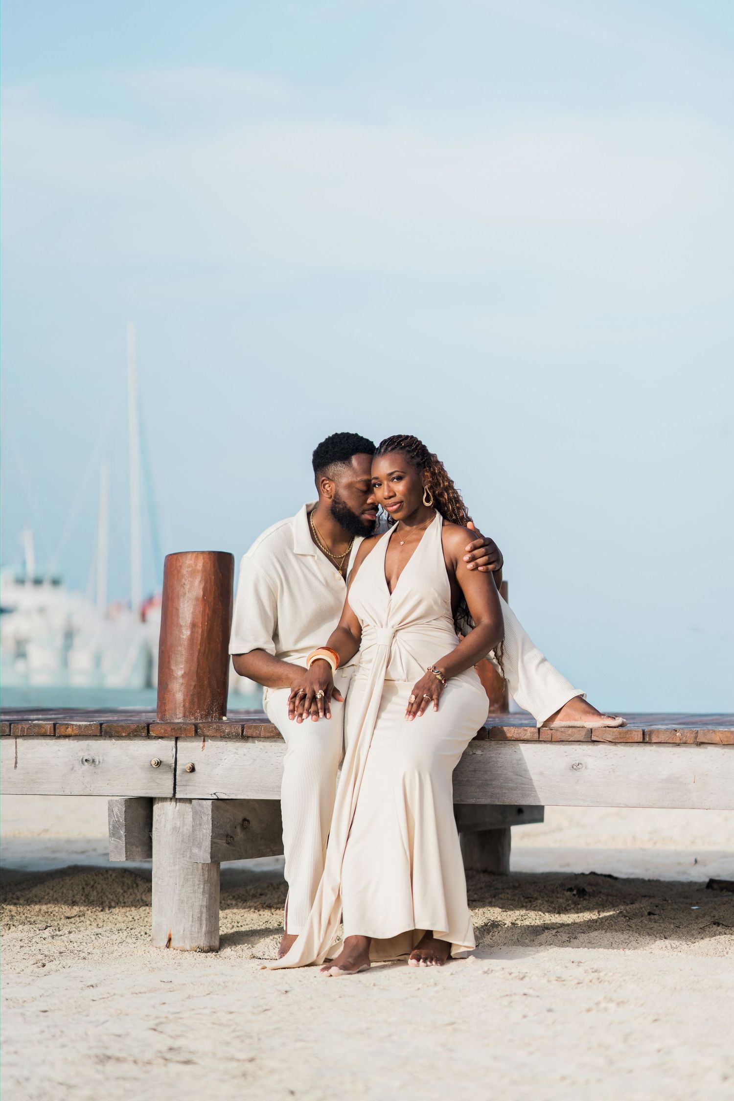 Romantic couple in cream attire sharing an intimate moment on a sandy beach bench.