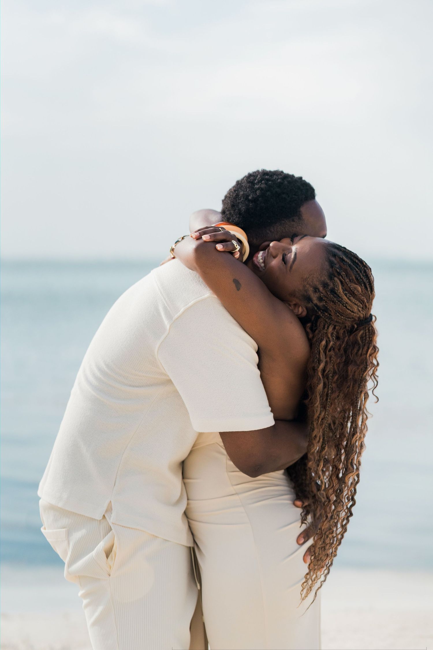 A couple sharing a romantic kiss on a sandy beach, both dressed in elegant cream outfits with the ocean in the background.