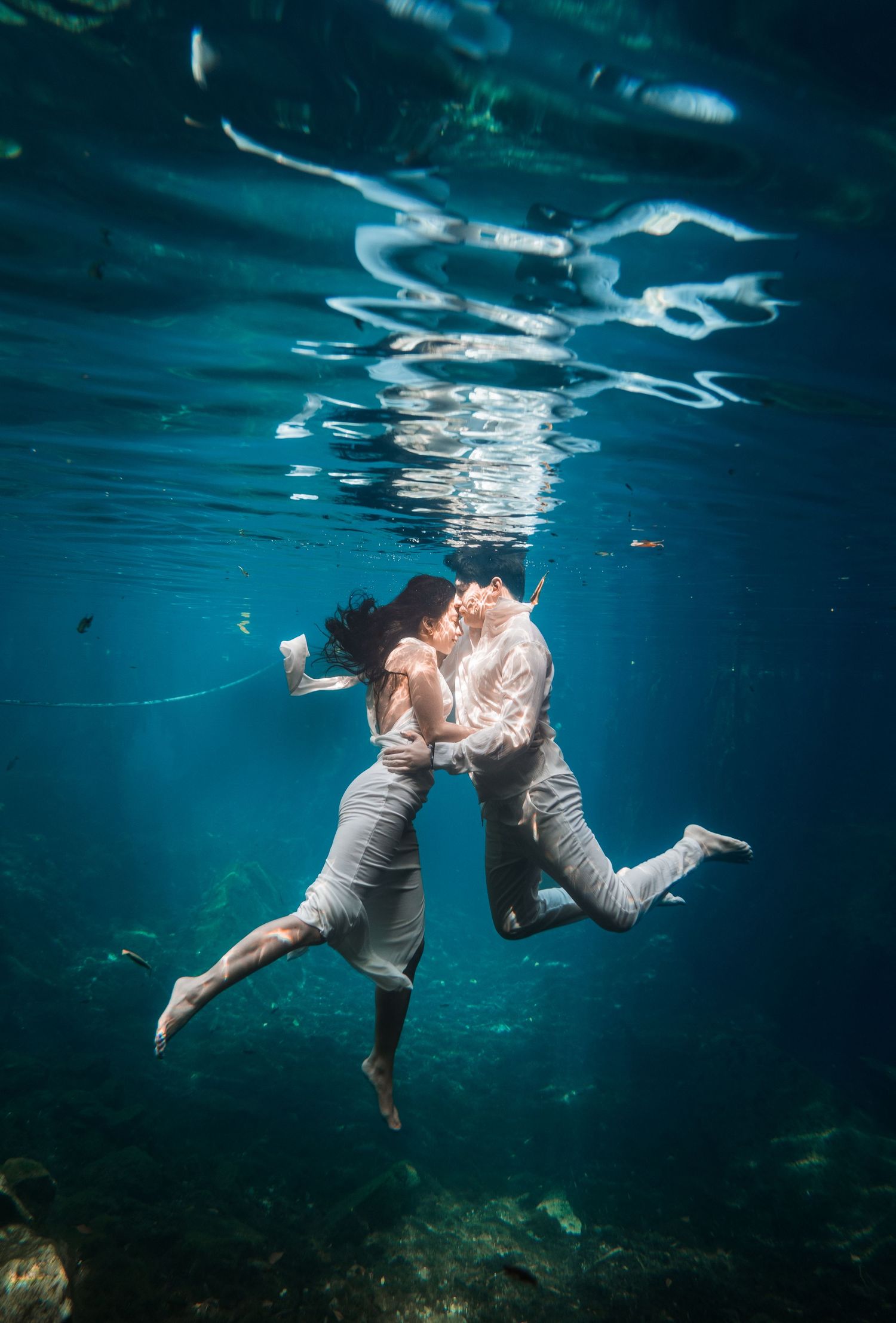 A couple shares a romantic underwater kiss, dressed in white, surrounded by clear blue water.
