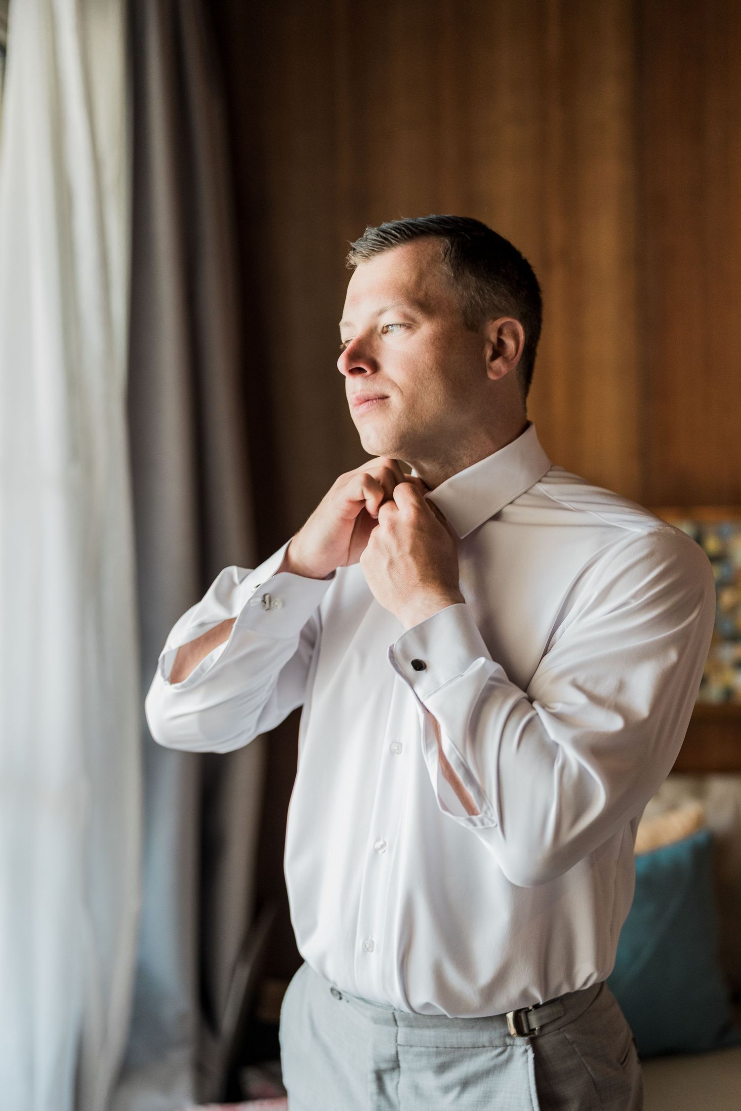 Man in white dress shirt adjusting his tie near a window, getting dressed for a formal occasion.