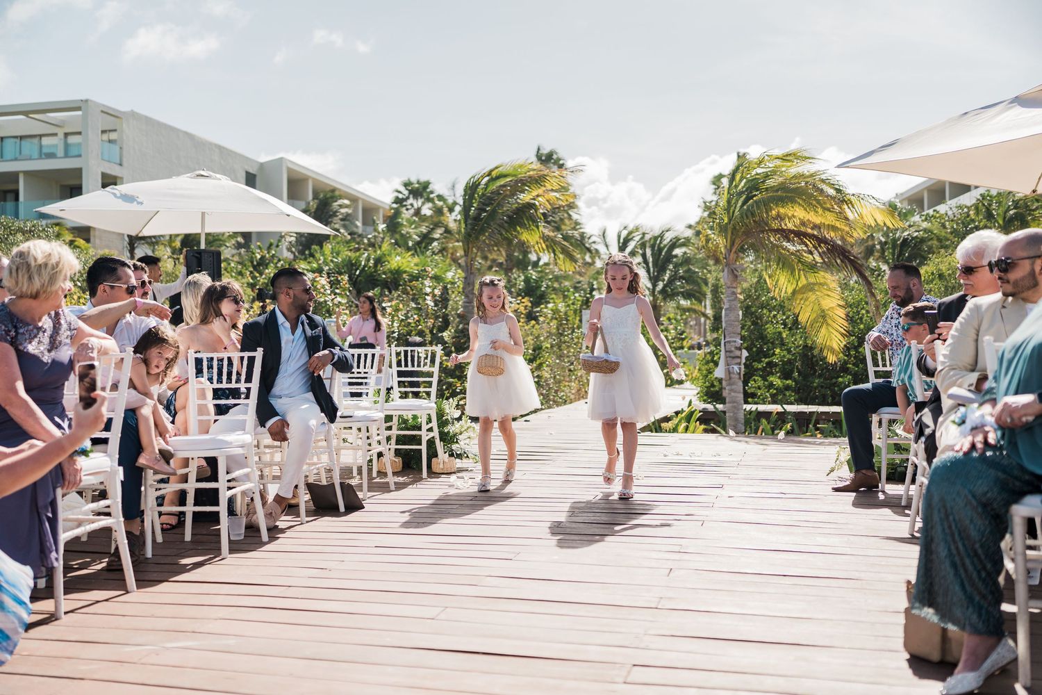 Two flower girls walk down an outdoor wedding aisle on a wooden deck surrounded by guests and tropical palm trees.