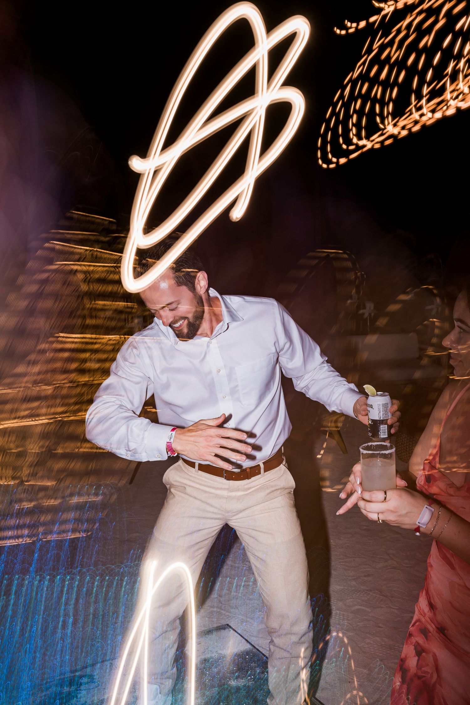 Man dancing at night party with light trails and bokeh effects in background, holding drink. Hard Rock Riviera Maya wedding photographer