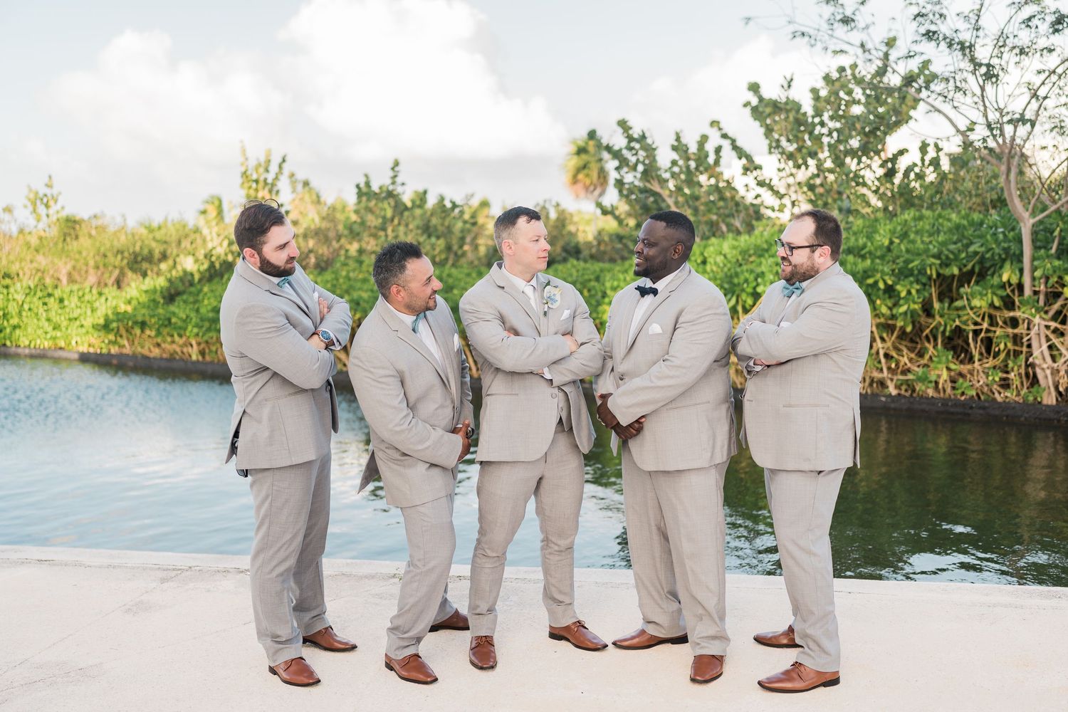 Five groomsmen in tan suits pose confidently by a waterway at an outdoor wedding venue.
