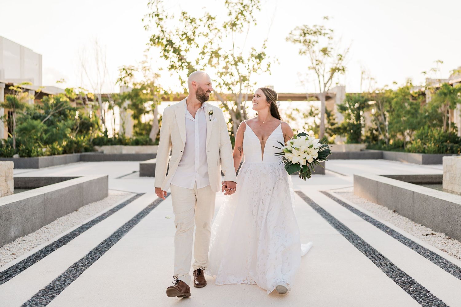 Bride and groom walk hand-in-hand in elegant white outdoor venue, she in lace gown, he in cream suit.