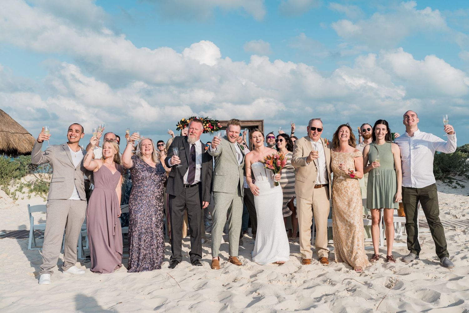 Wedding party celebrating on a sandy beach with bride and groom surrounded by guests in formal attire under a blue sky.