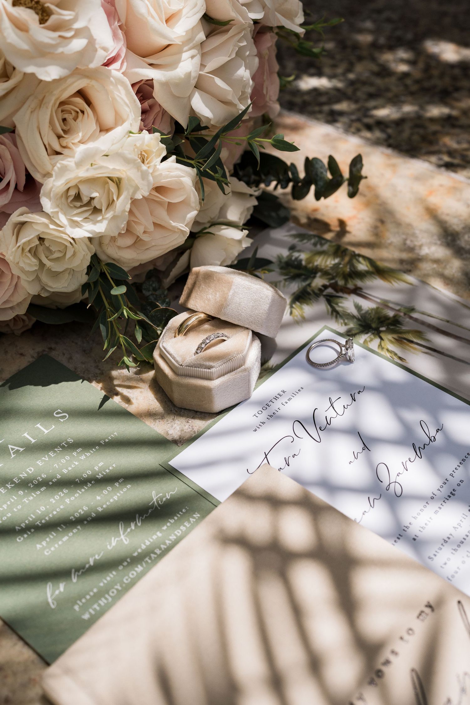 Elegant wedding flat lay with cream roses bouquet, ring box, gold bands, and sage green calligraphy invitations in soft natural light.