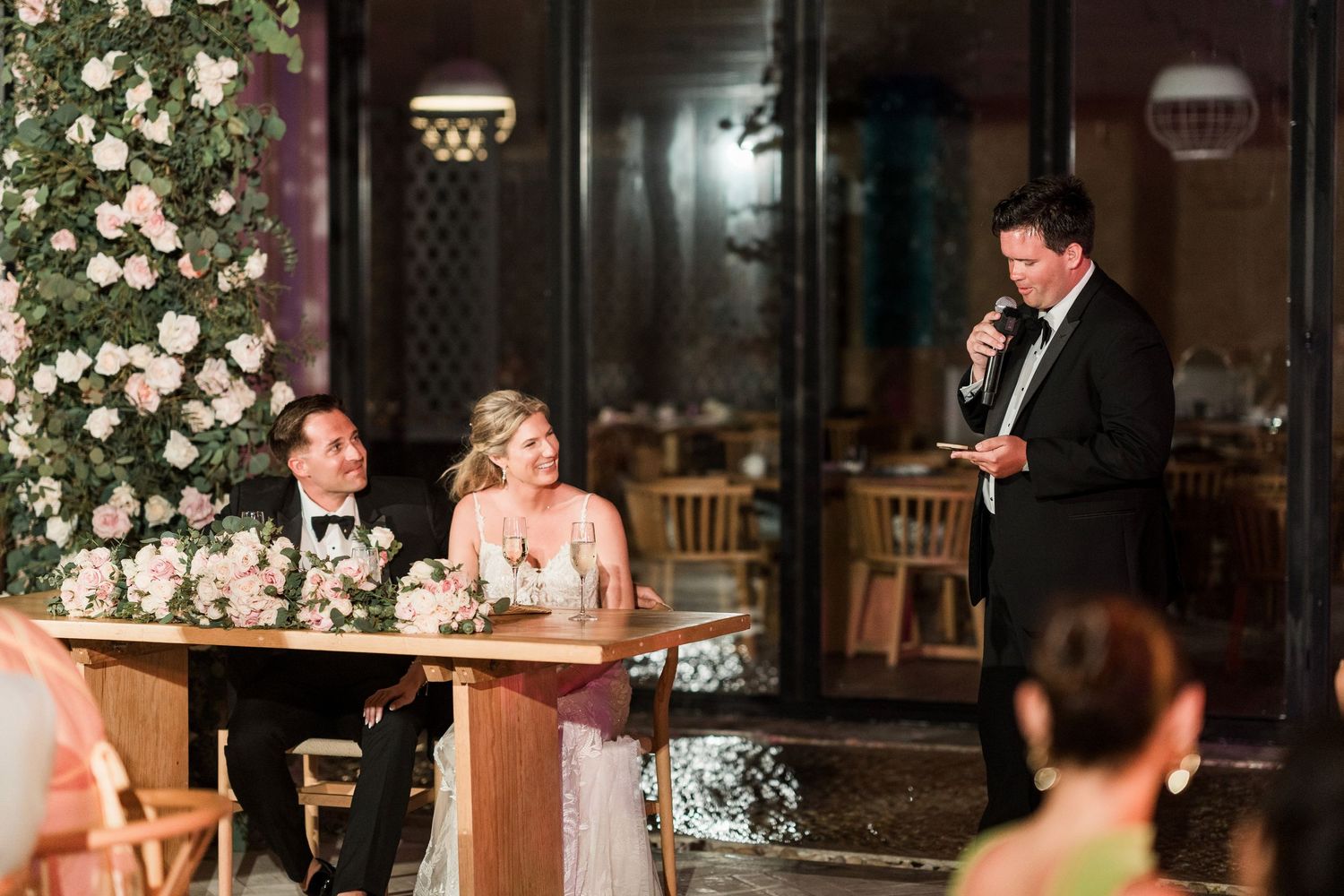 Groom gives wedding speech at elegant reception while bride and groom sit at floral-adorned sweetheart table.