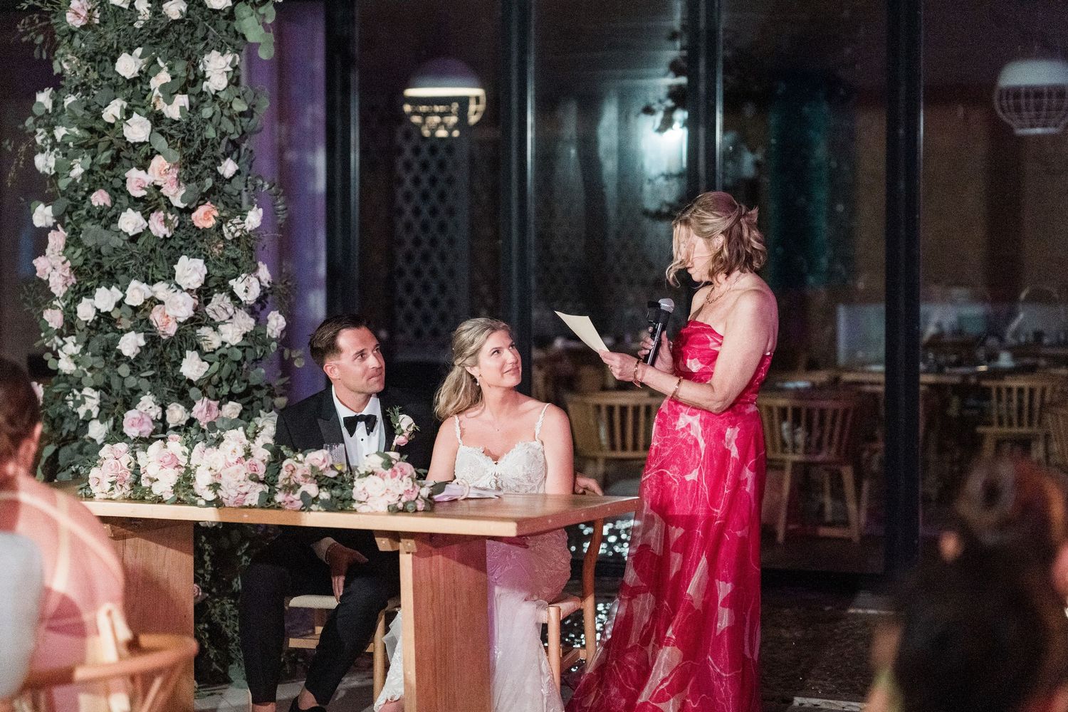 Woman in red gown gives wedding speech to bride and groom seated at floral-decorated sweetheart table.