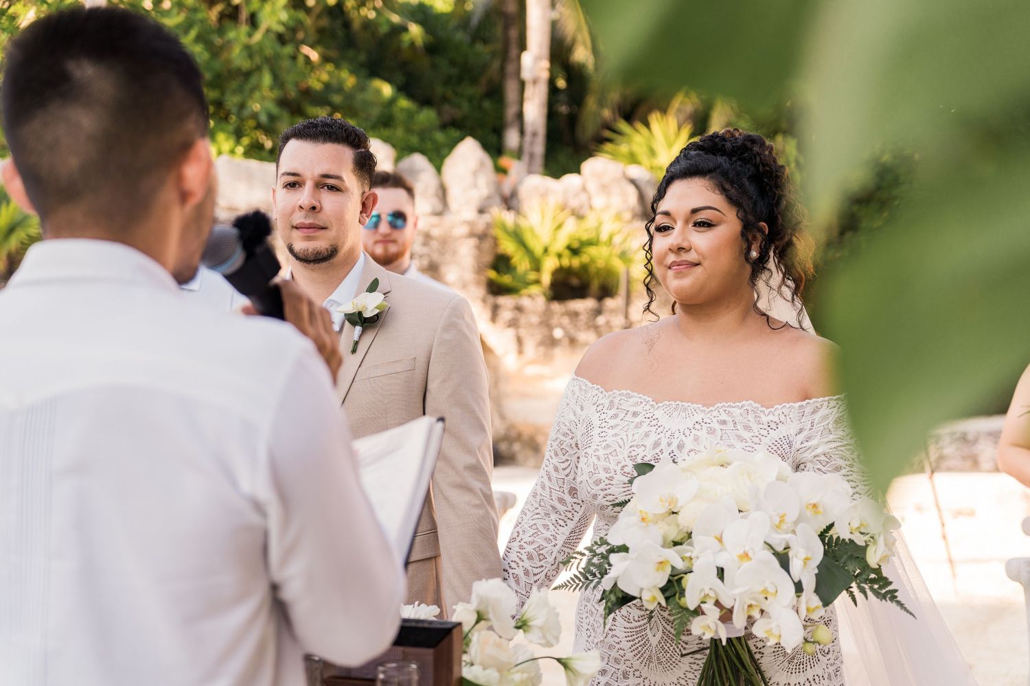 Xcaret Arte Wedding - Bride in beaded off-shoulder gown holding white bouquet during outdoor wedding ceremony with groom in tan suit.