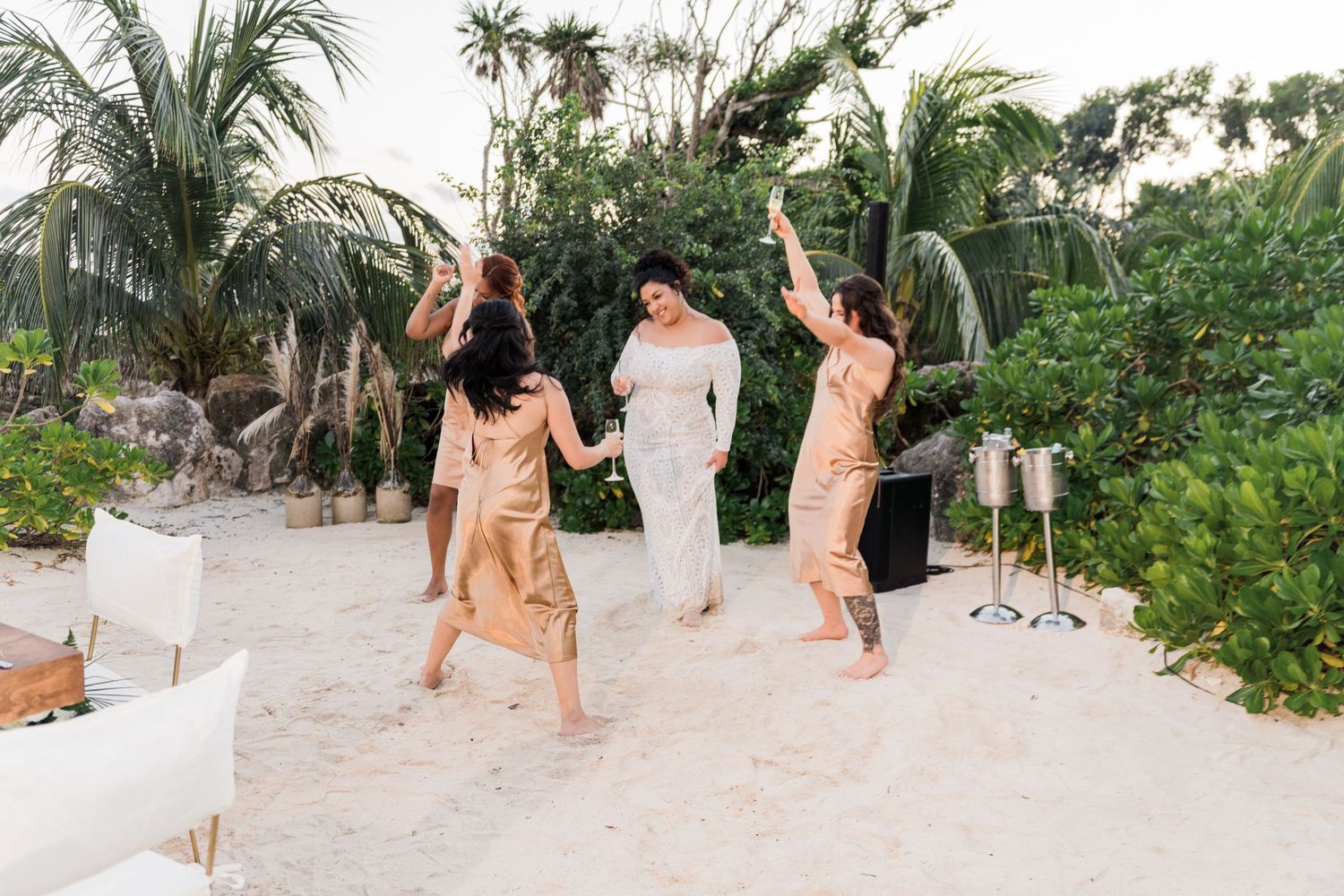 Xcaret Arte Wedding - Bride in white gown dancing with bridesmaids in gold dresses on sandy beach surrounded by tropical greenery.