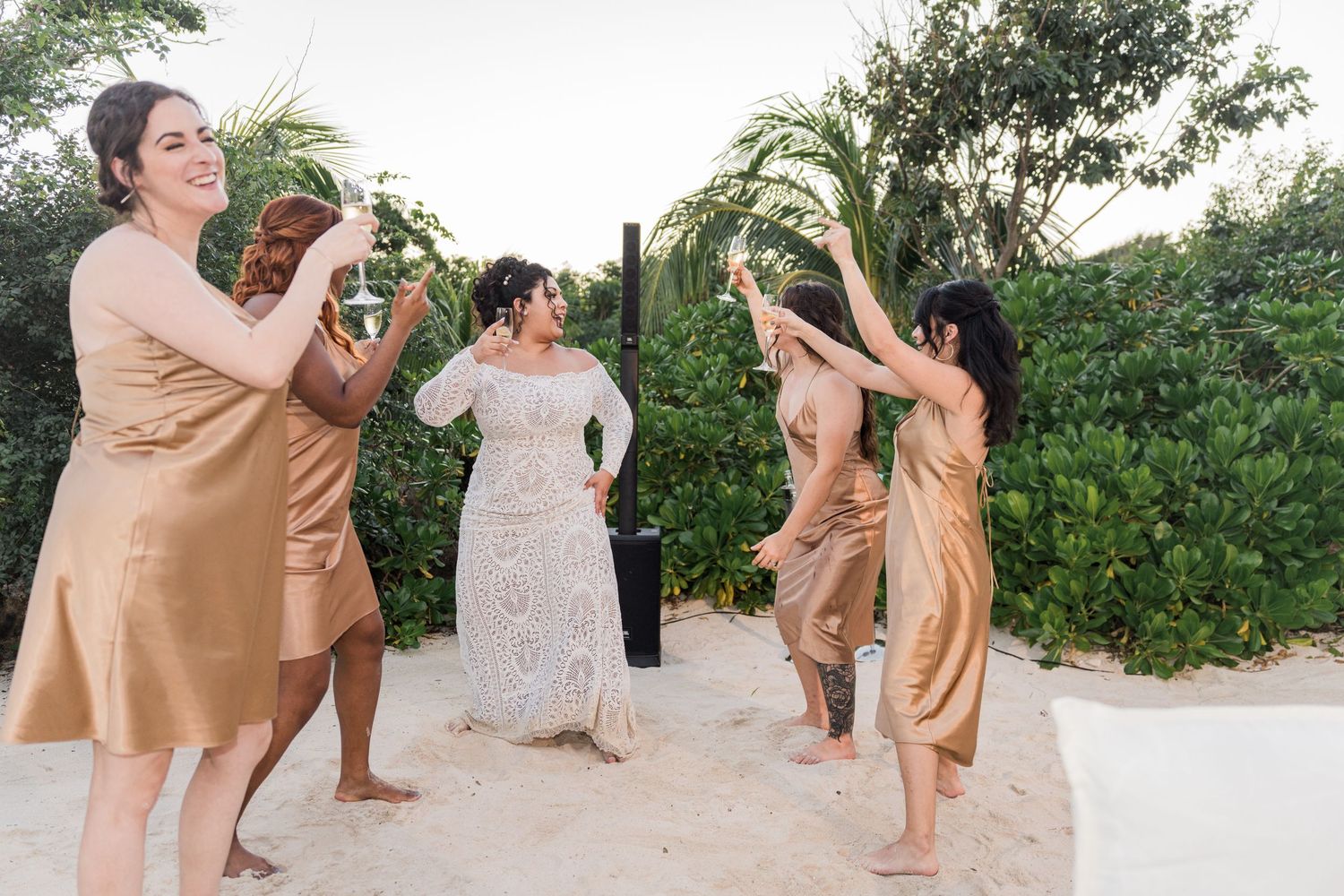 Xcaret Arte Wedding - Bride in white gown celebrates with bridesmaids in gold dresses on sandy beach surrounded by tropical greenery.