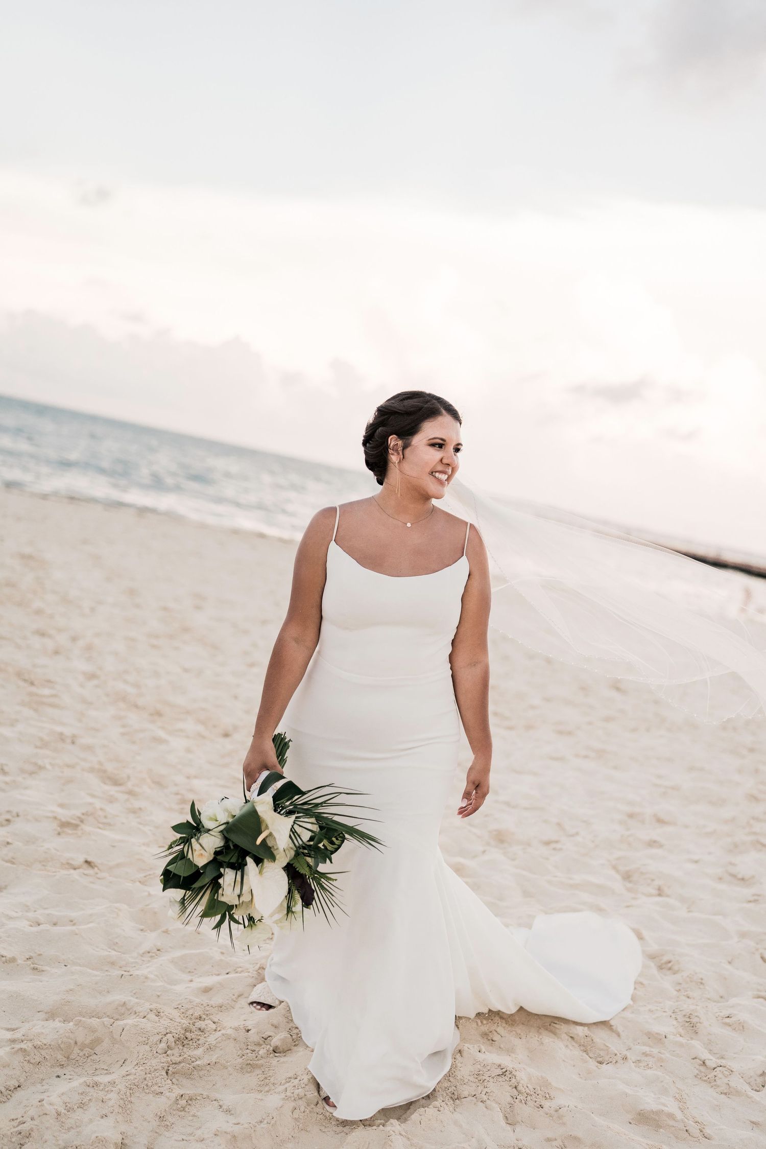 Bride in white spaghetti strap gown holding floral bouquet smiling on sandy beach at sunset.