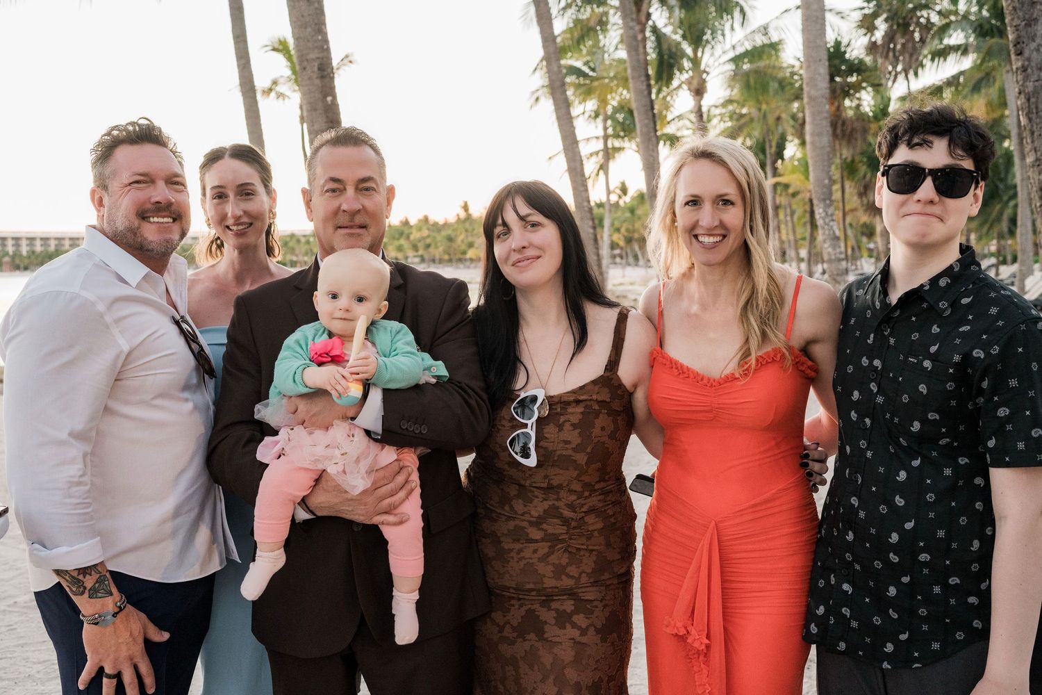 Group of seven people posing together outdoors near palm trees, one holding a baby in pink, dressed in semi-formal attire.