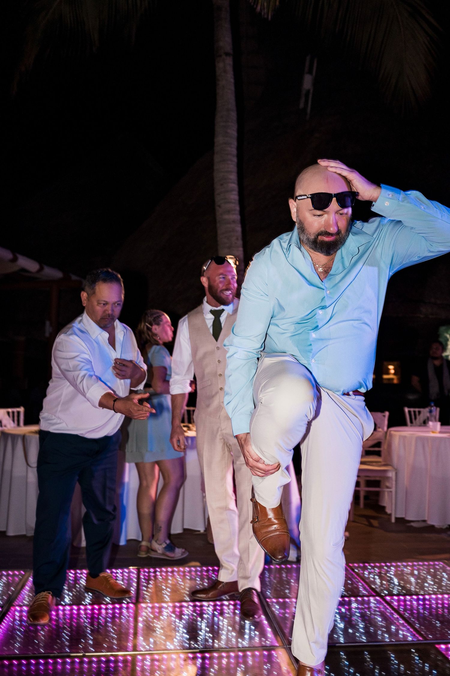 Bald man in sunglasses dancing energetically on illuminated dance floor at outdoor wedding reception at night.