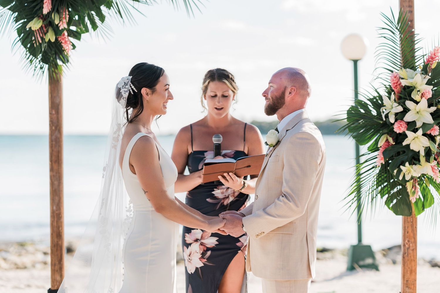 Bride and groom exchange vows on a tropical beach with a floral arch, officiated by a woman in a black dress.