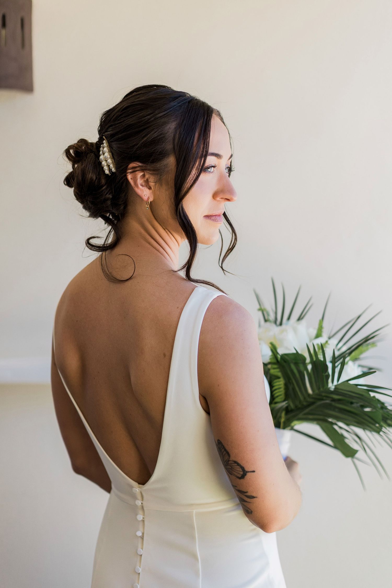 Bride in backless white gown holds bouquet, showing elegant updo and tattoo.