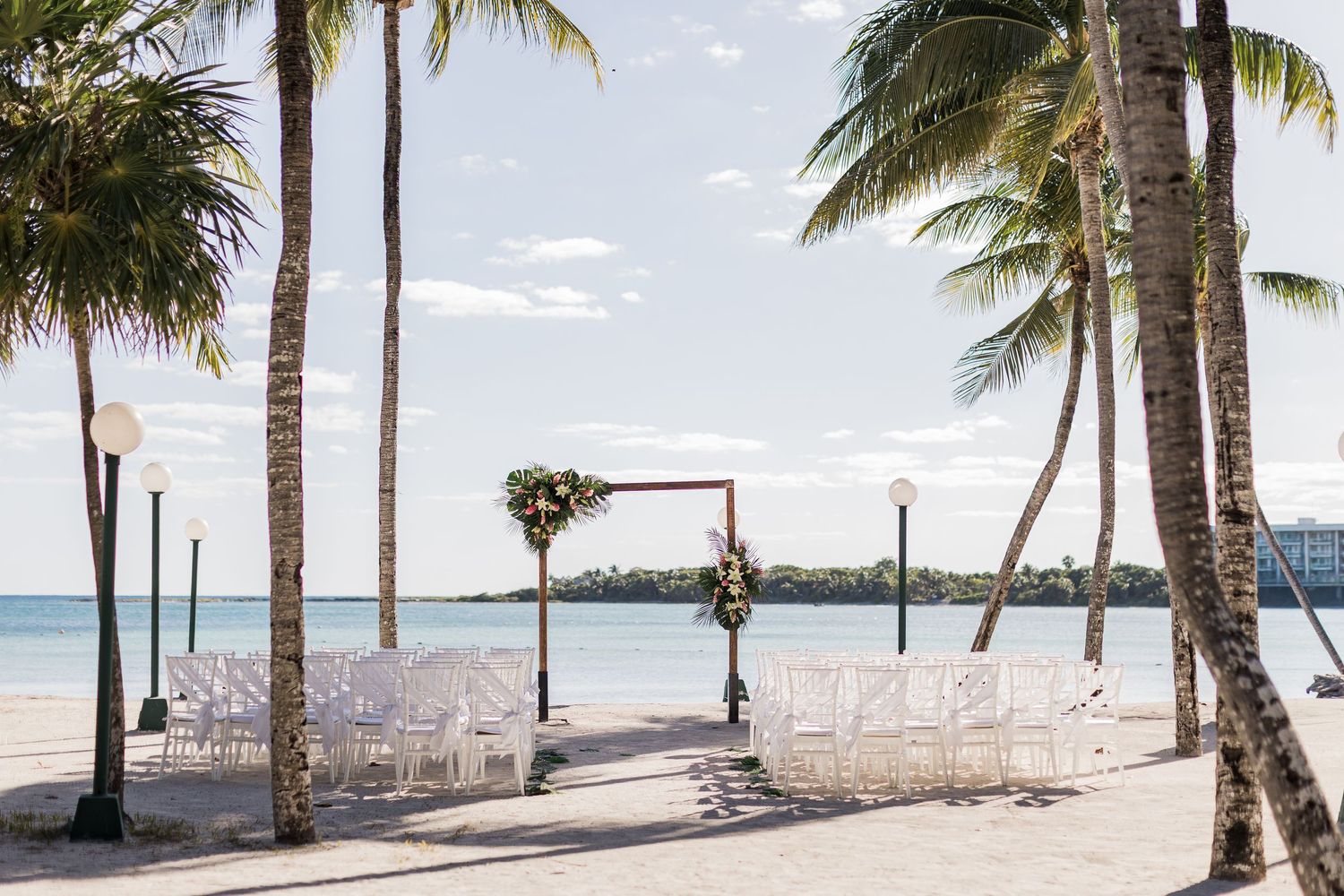 Outdoor beach wedding ceremony setup with white chairs, floral arch, and palm trees overlooking a calm turquoise ocean.