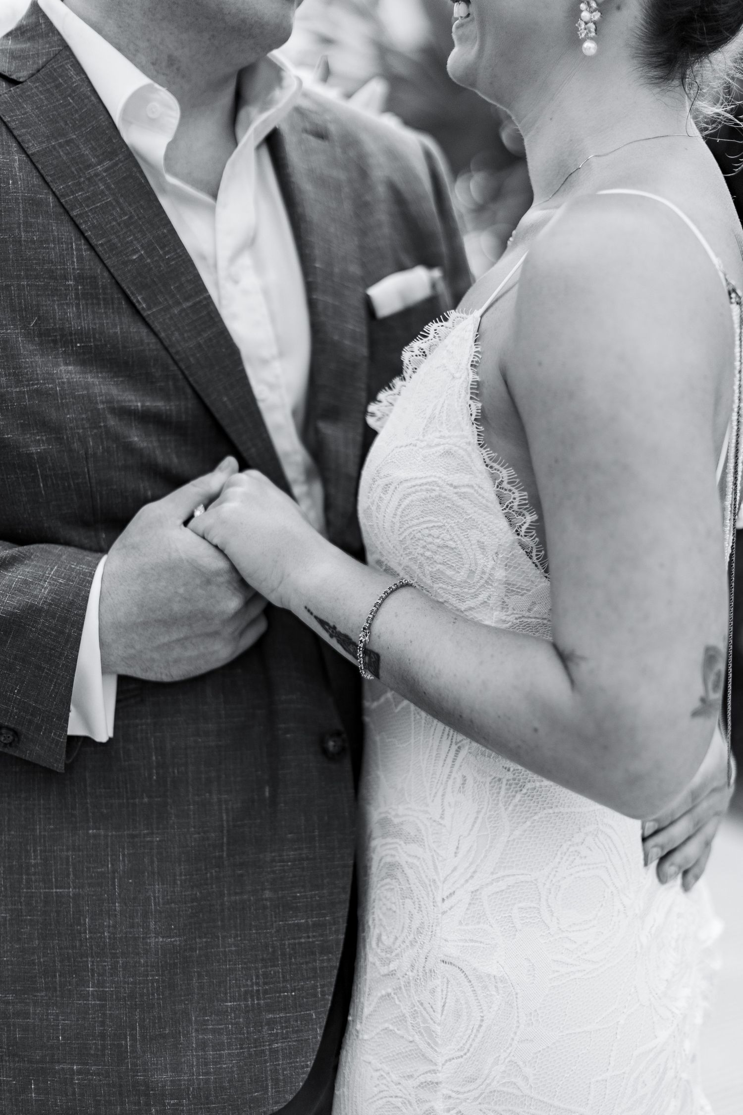 Black and white photo of bride and groom holding hands, bride in lace gown, groom in suit at wedding ceremony.