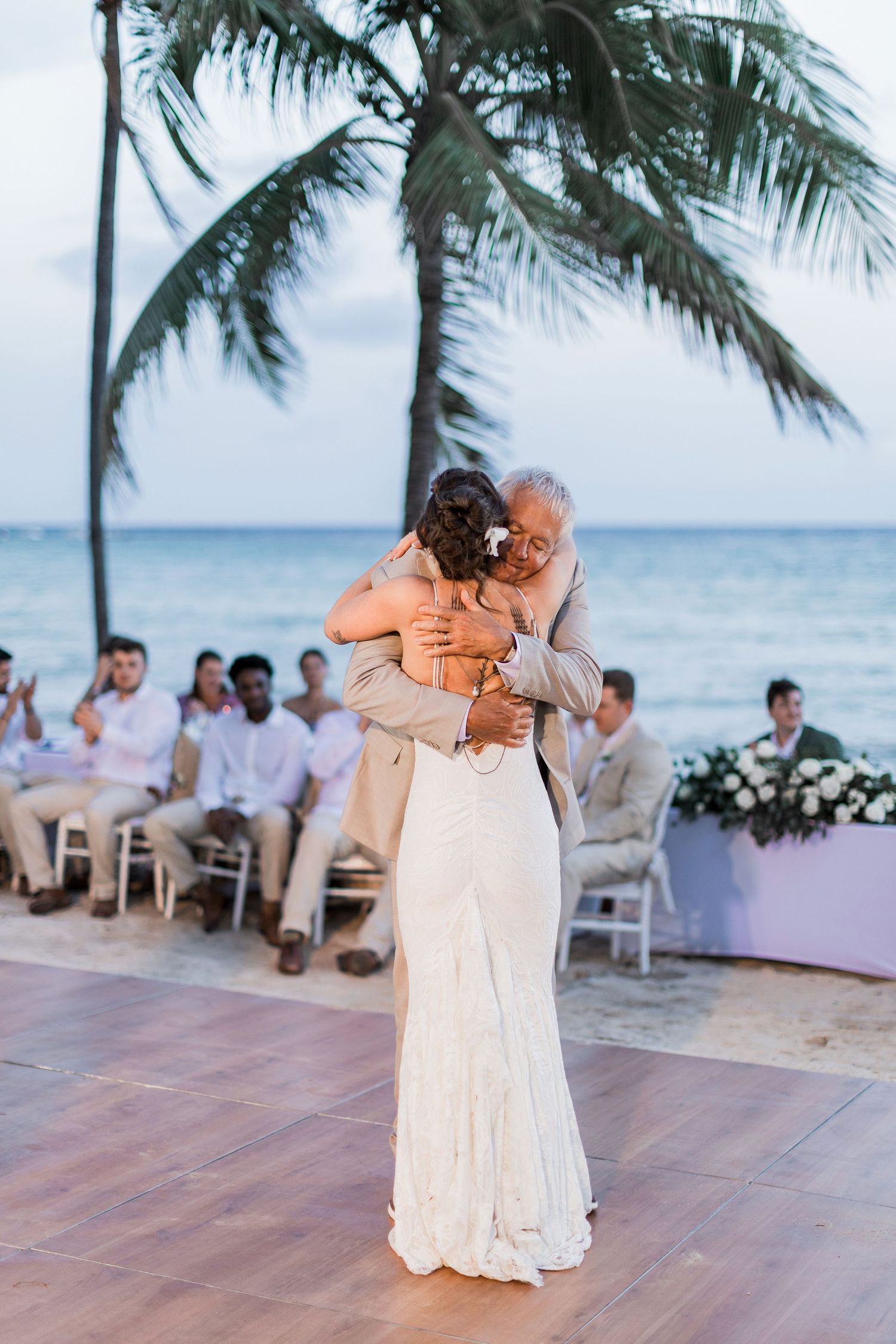 Bride and older man share emotional first dance at beach wedding with palm trees and ocean backdrop.