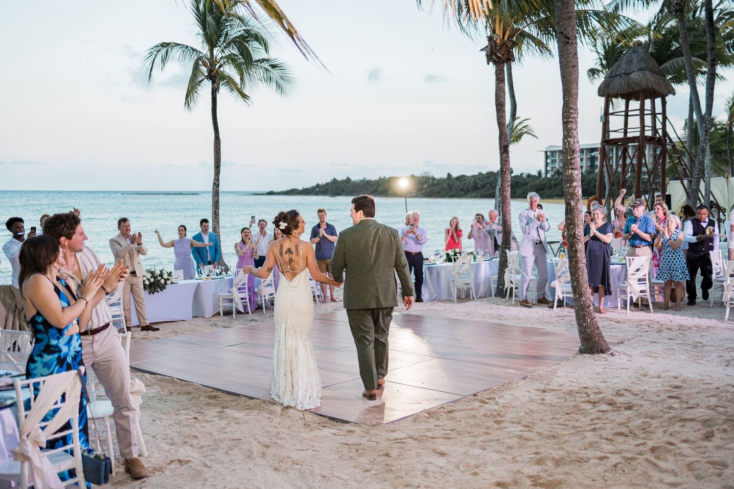 Bride and groom walk hand-in-hand on sandy beach wedding venue with guests applauding, palm trees, and ocean backdrop.
