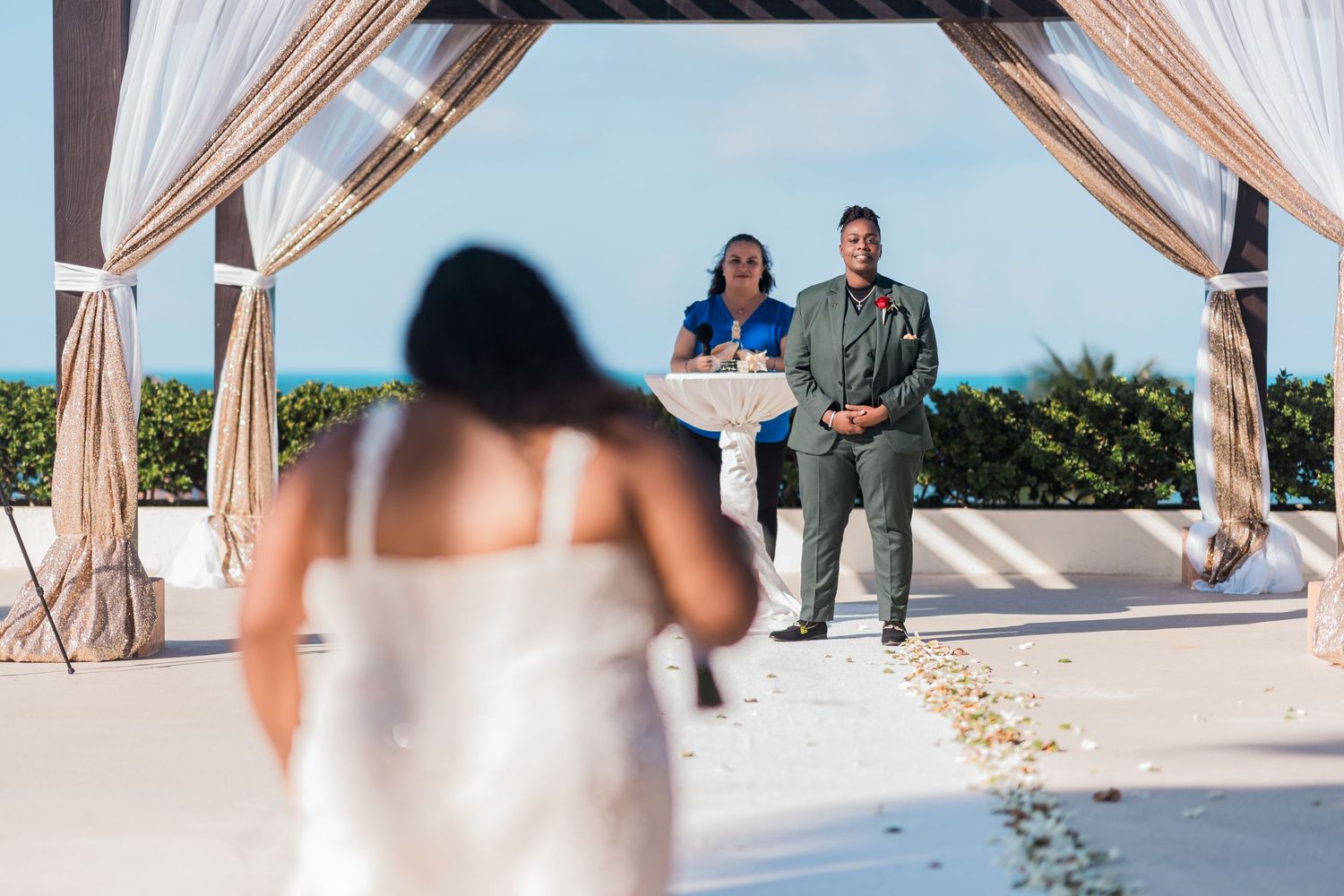 Bride walking down the aisle toward groom and officiant at an outdoor beach wedding ceremony.
